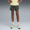 Op grijs, een model poseert in een gele top, grijze short met logo, sokken en hardloopschoenen.