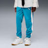 Tegen een grijze achtergrond draagt een model een turquoise broek, witte sneakers en een wit overhemd met een zwarte das.