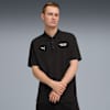 Vor grauem Hintergrund posiert ein Model in schwarzem Poloshirt mit Logos und dunkler Hose.