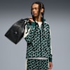 Sur fond gris, un mannequin en survêtement vert à motifs porte un sac noir.