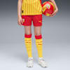 Su sfondo grigio, modello tiene un pallone indossando kit calcio giallo e rosso e scarpe da ginnastica.