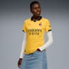 Tegen een grijze achtergrond poseert een model in een geel voetbalshirt, shirt met lange mouwen en denim rok.