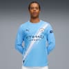 Against grey, a model wears a light blue, long-sleeved sports shirt, posing with arms behind.