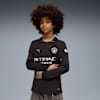 Op grijs poseert een model in een zwart Manchester City shirt met lange mouwen en grijze jeans.