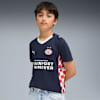 Un modèle se tient devant un fond gris, portant un t-shirt de foot PSV bleu foncé et un jean bleu clair.