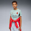 Sur un fond gris, un enfant porte un t-shirt à motifs avec une veste rouge nouée autour de lui.