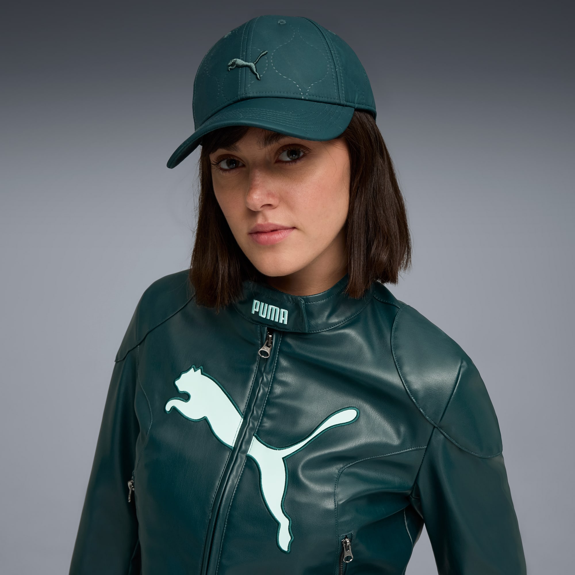 Un modèle pose sur fond gris, portant une casquette et une veste verte de marque.