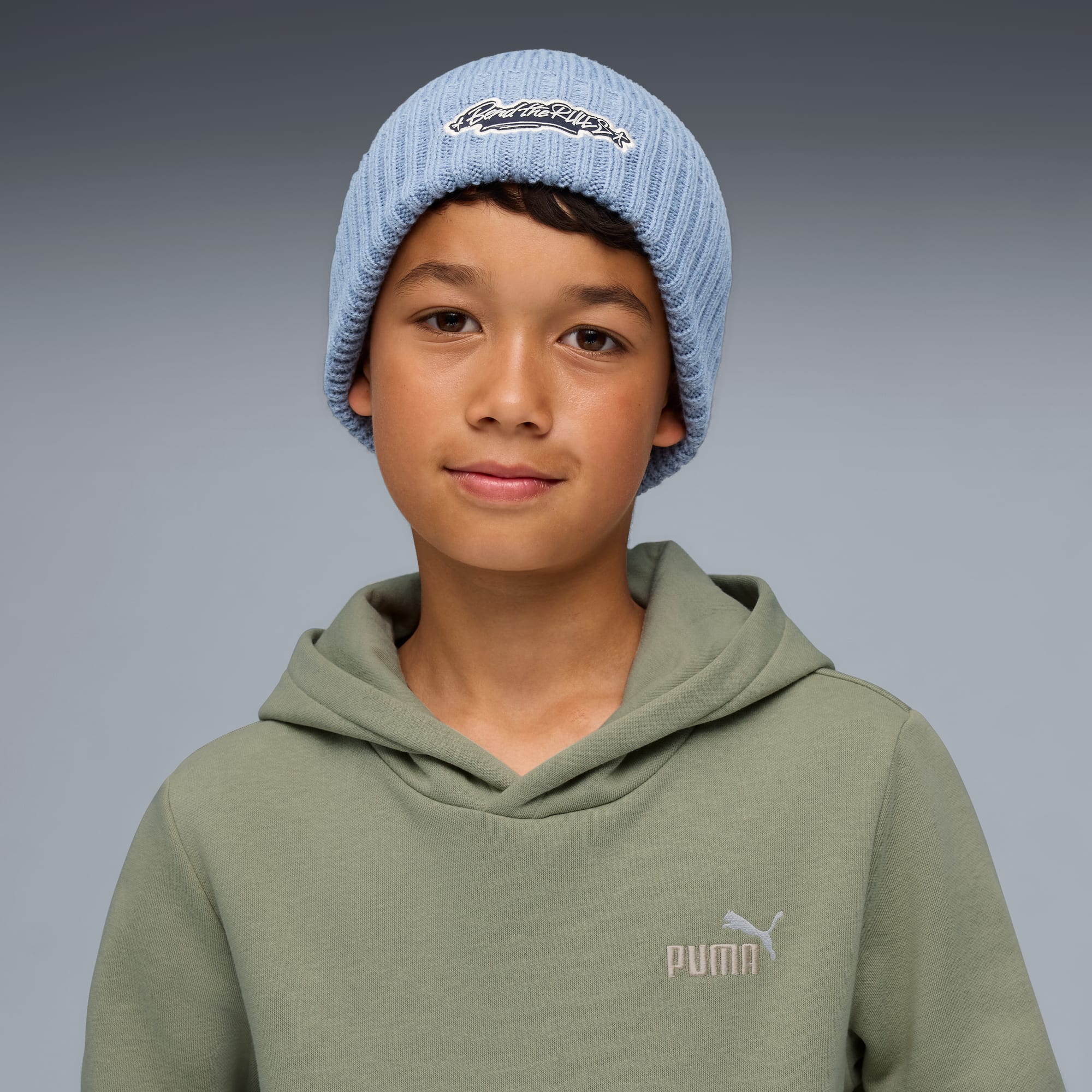 Een model voor een grijze achtergrond met een blauwe muts en een olijfgroene hoodie.