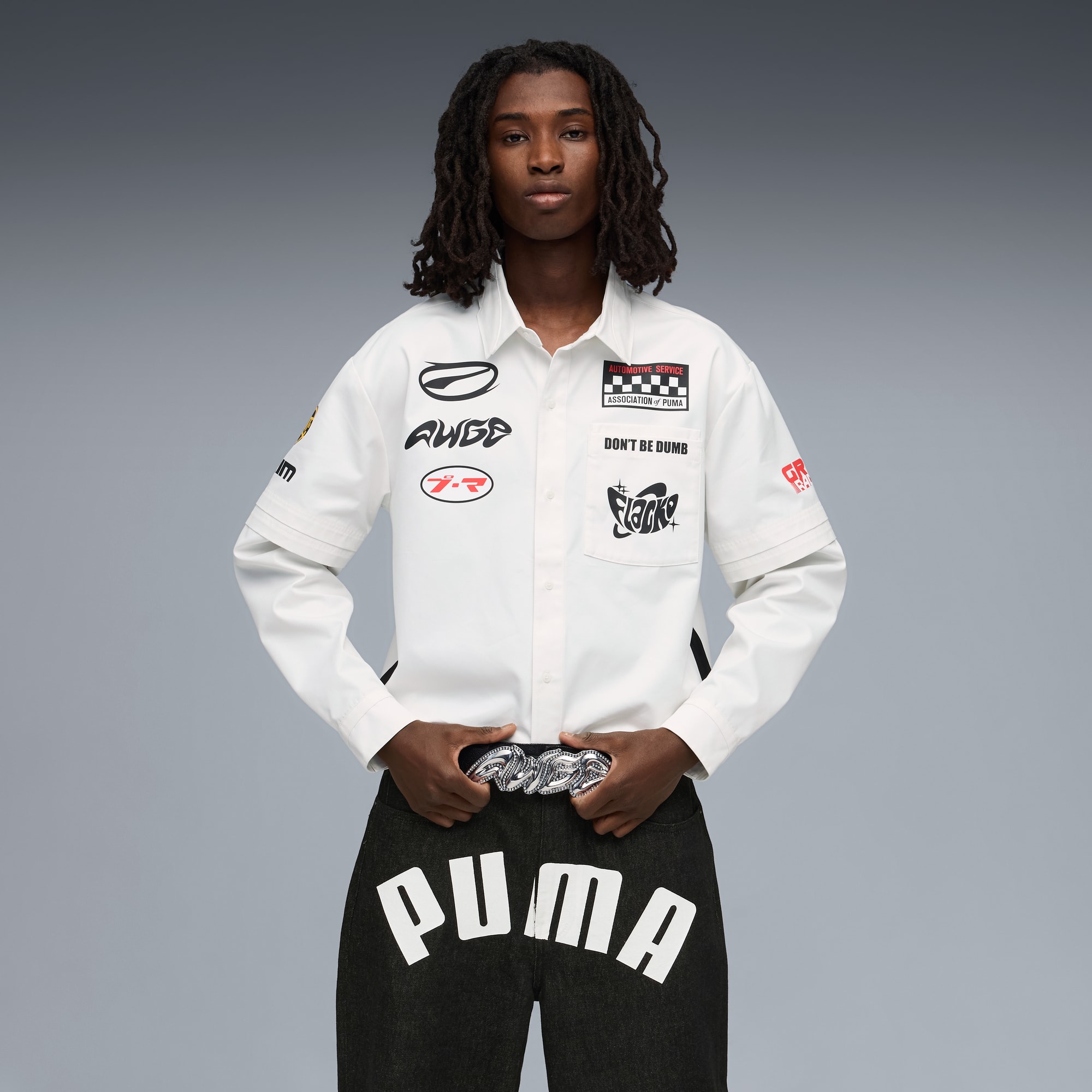 Een model poseert tegen grijs, draagt een wit overhemd met emblemen en een donkere PUMA broek.