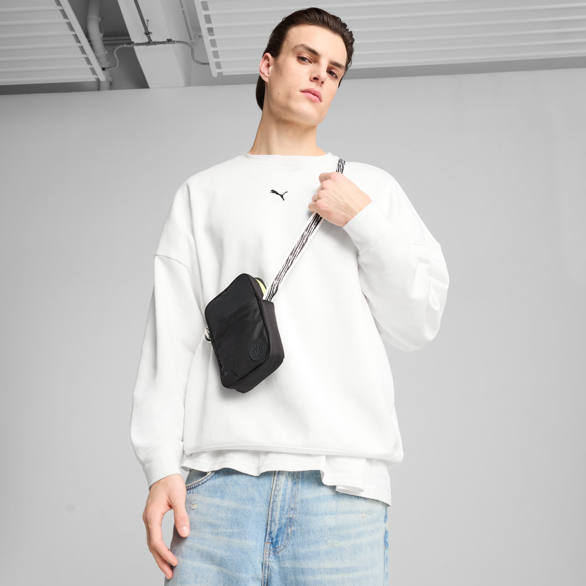 Sur fond gris, un mannequin pose avec un sac bandoulière noir et un pull blanc.
