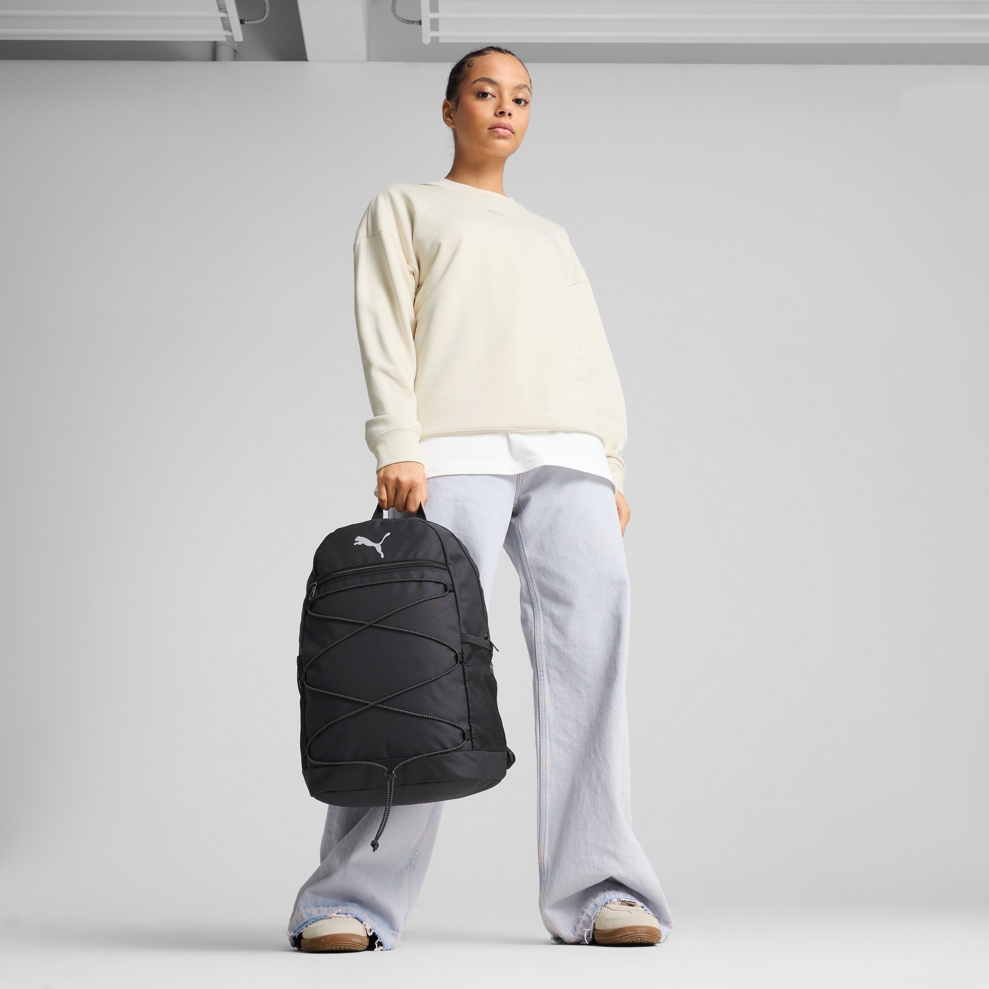 Su sfondo grigio, una modella tiene uno zaino nero, indossando top beige e jeans blu.