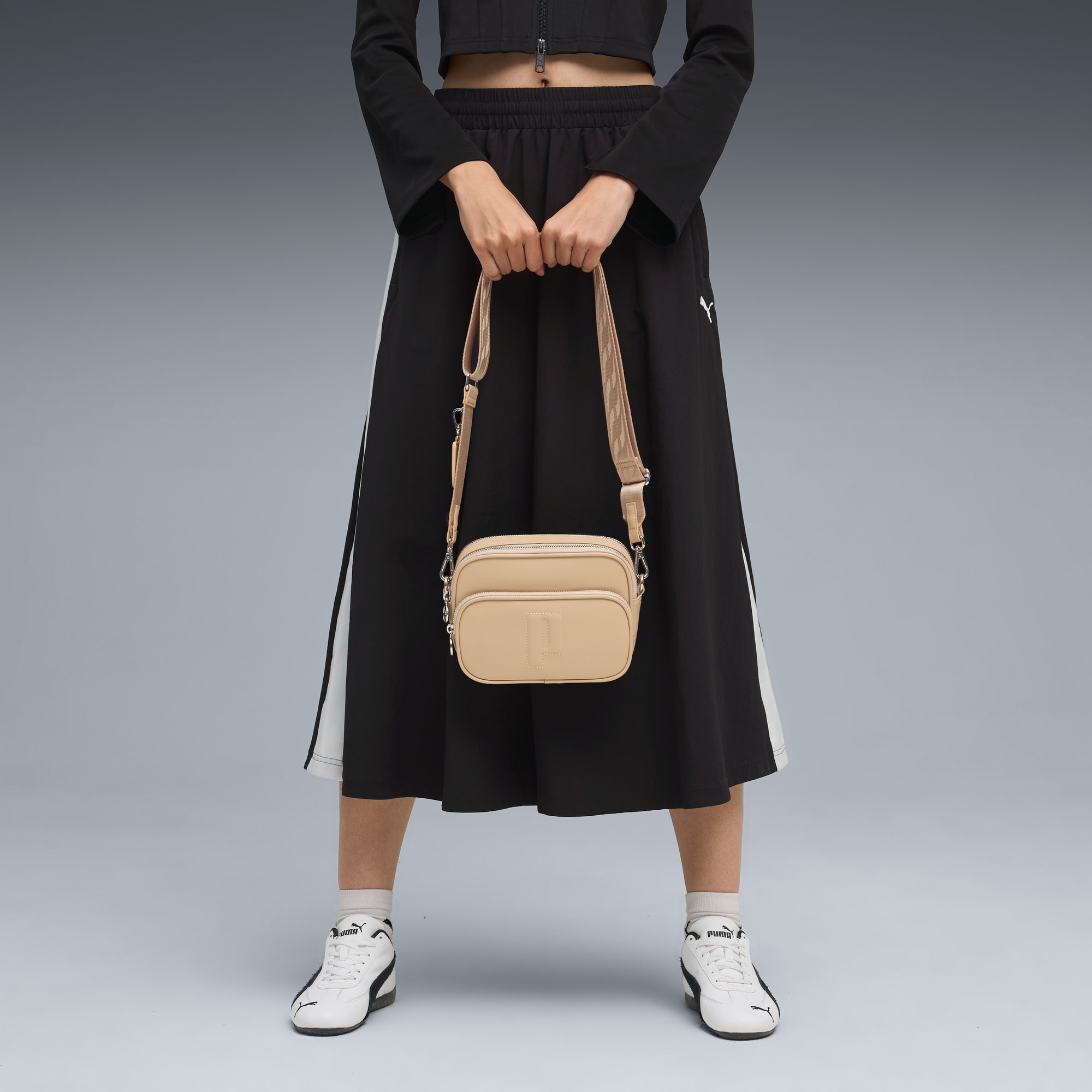 Vor grauem Grund hält ein Model eine beige Tasche, trägt Top, Rock und Sneaker.