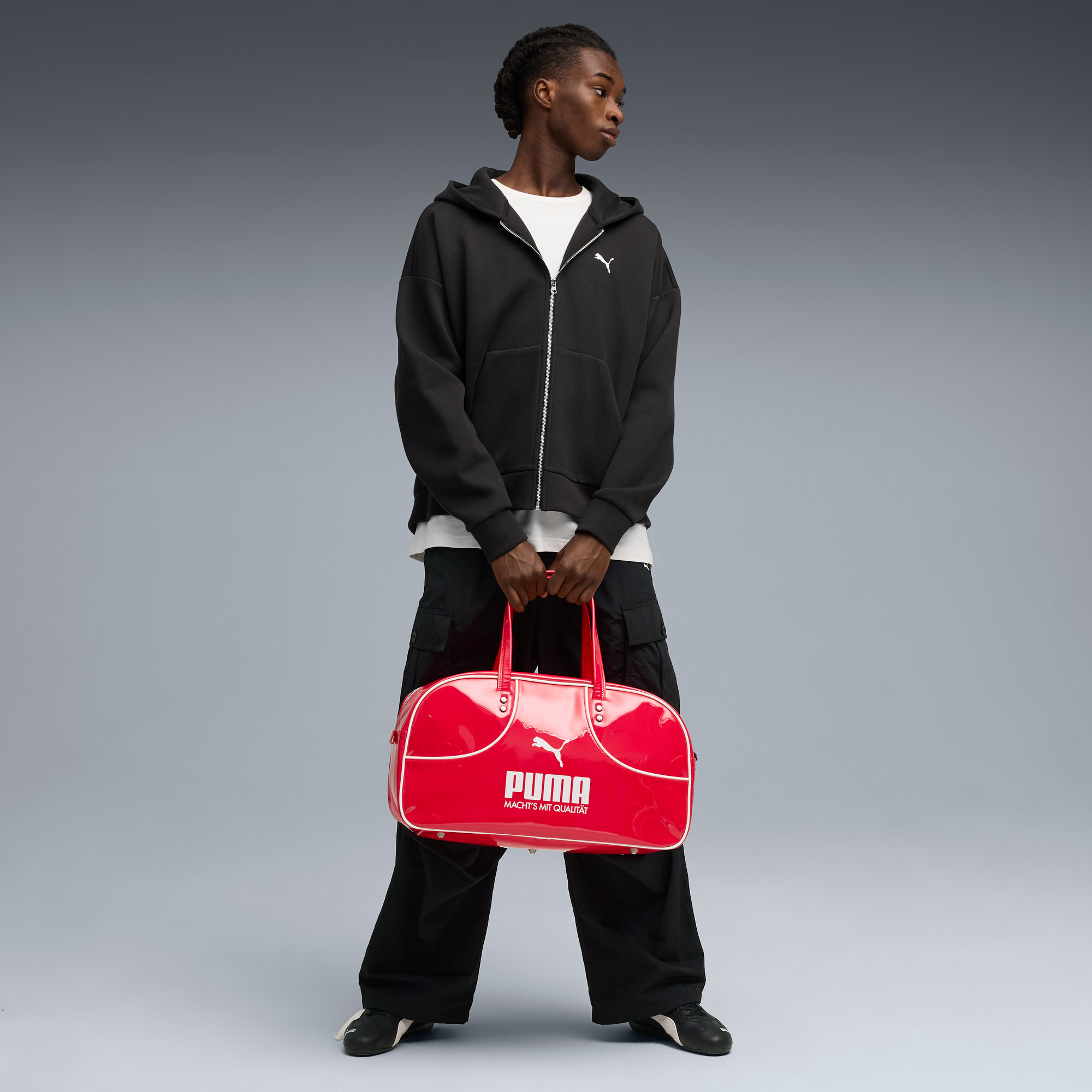 Model vor grauem Hintergrund hält rote Puma-Tasche, trägt schwarze Jacke mit Kapuze und Hose.