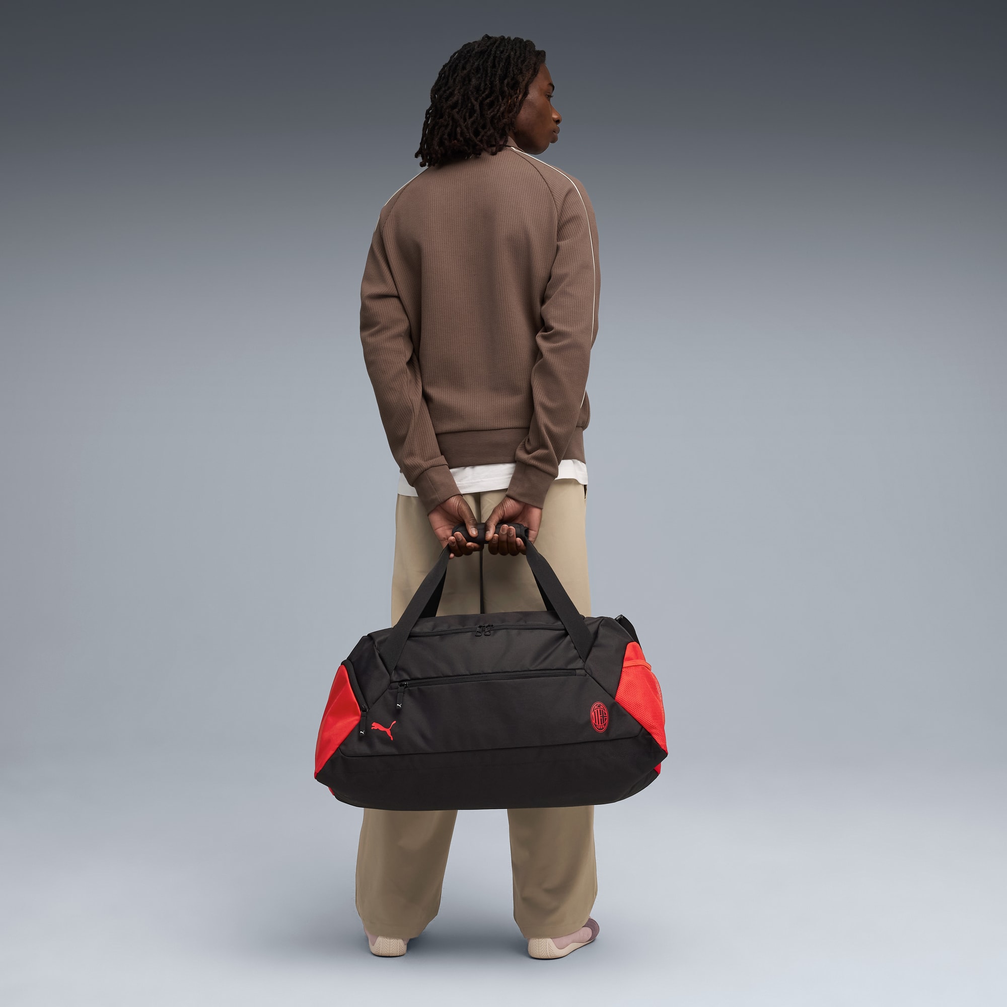 Vor grauem Hintergrund hält ein Model eine schwarz-rote Reisetasche und wendet sich von der Kamera ab.
