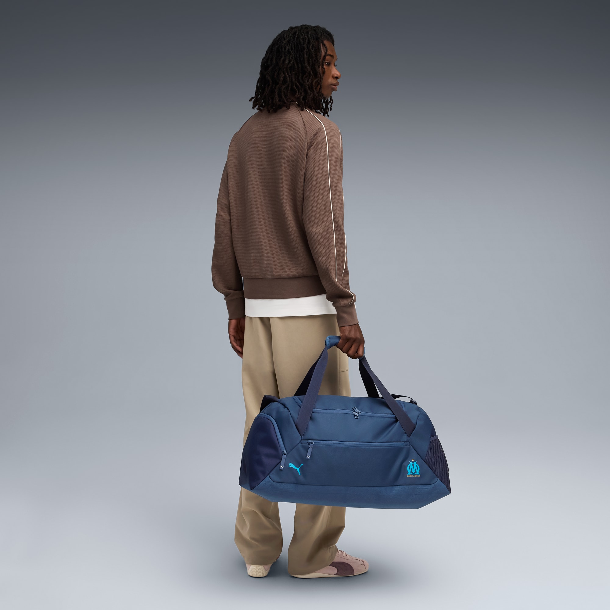 Su sfondo grigio, un modello tiene una borsa da palestra blu, con giacca marrone e pantaloni beige.