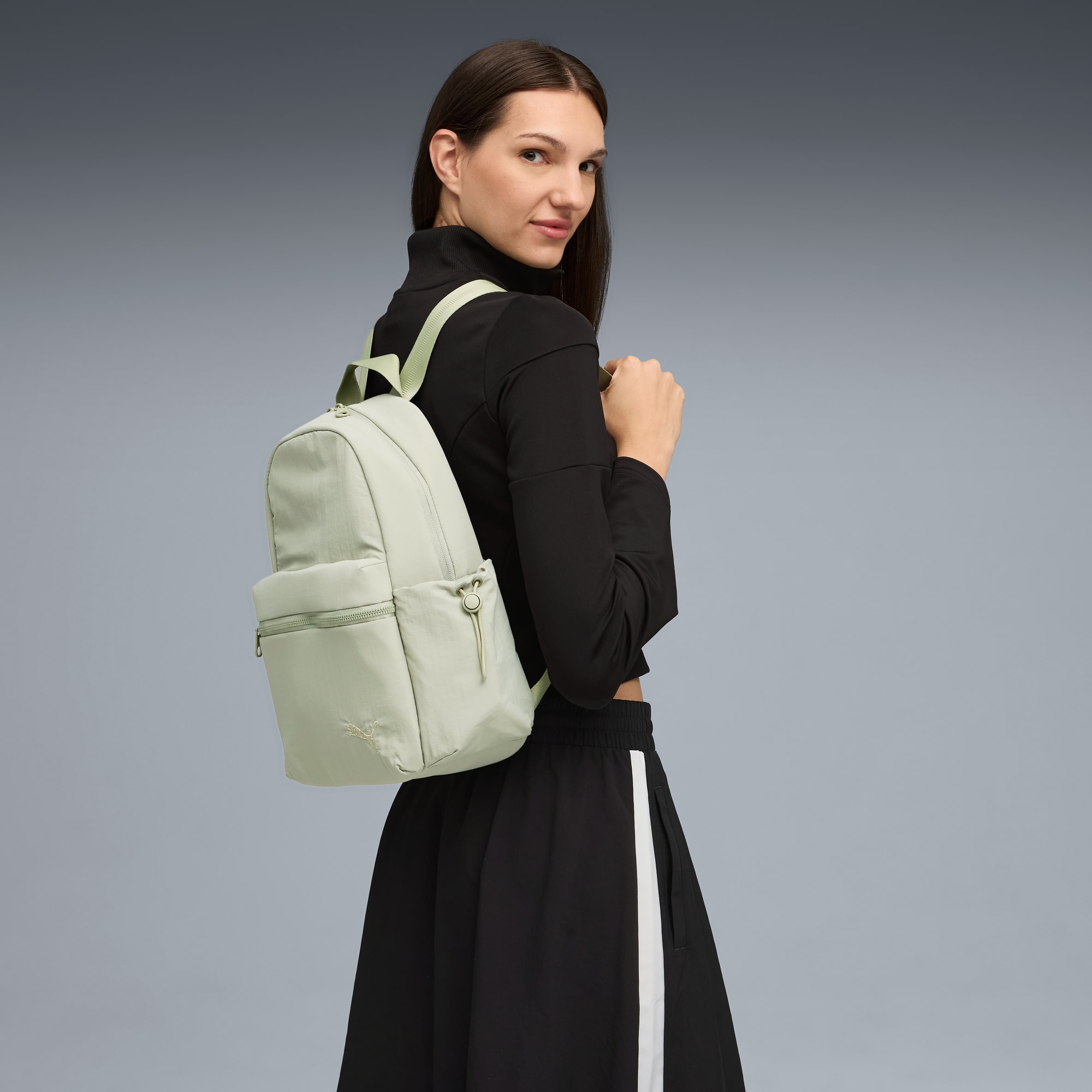 Vor grauem Hintergrund blickt ein Model zurück, mit schwarzem Oberteil, Rock und hellgrünem Rucksack.