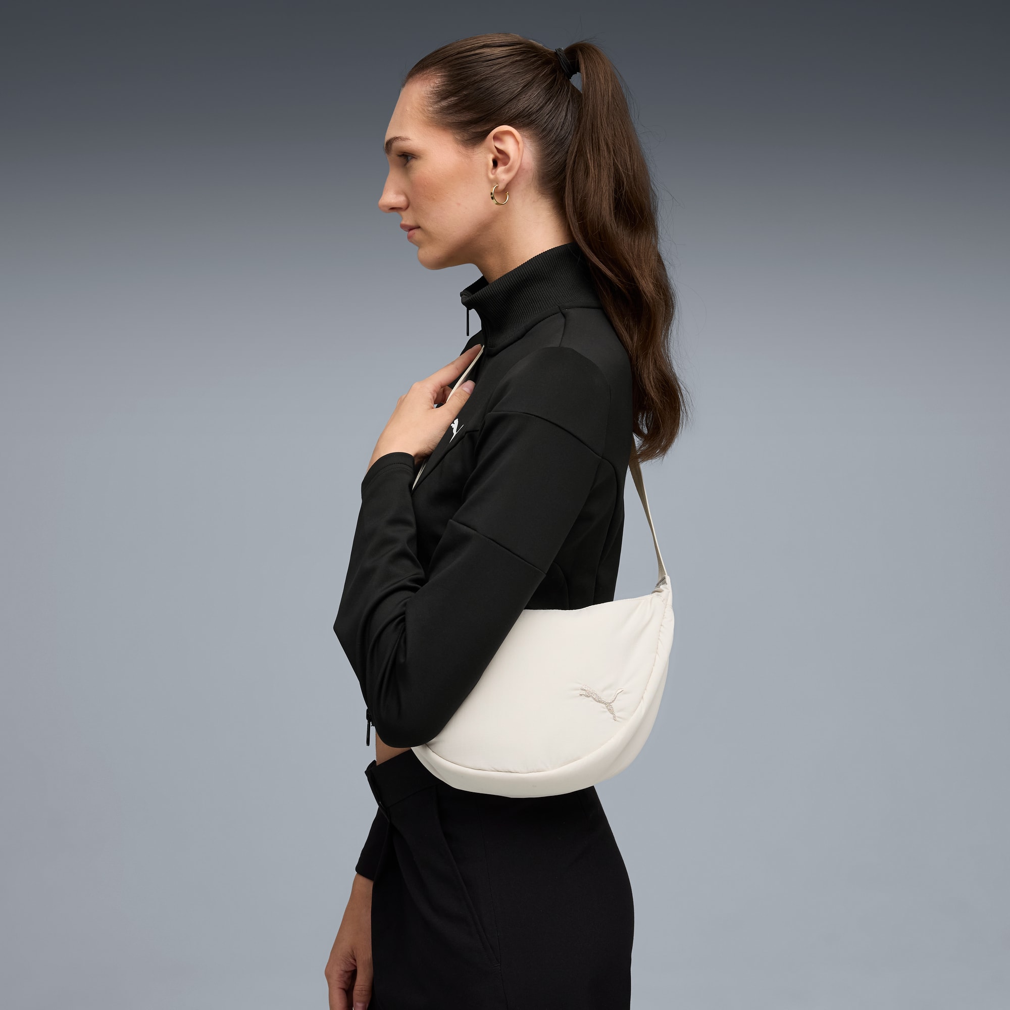 Sur fond gris, un mannequin pose en tenue noire avec un sac blanc en forme de croissant.