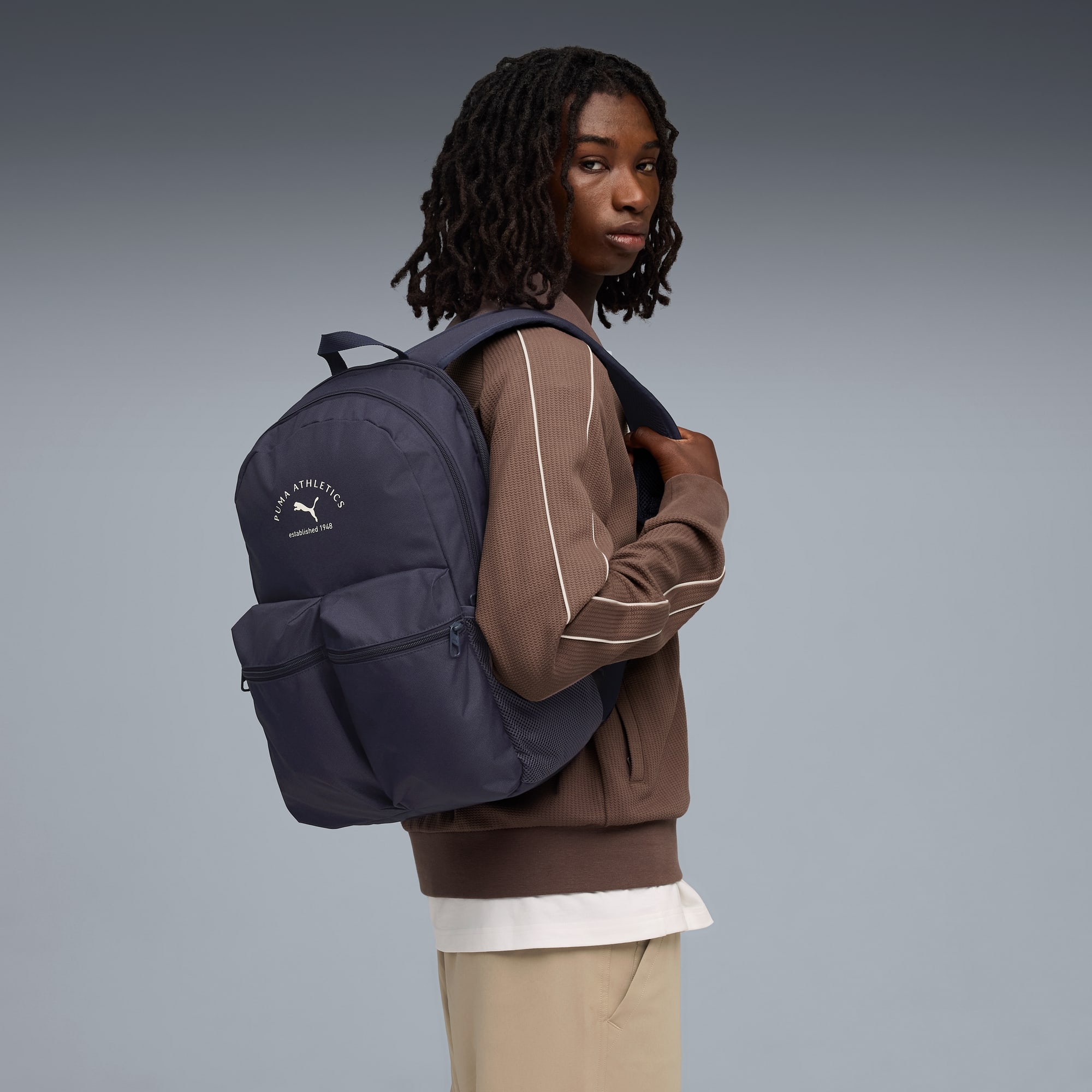 Sur fond gris, un mannequin regarde en arrière, portant un haut marron et un sac Puma marine.