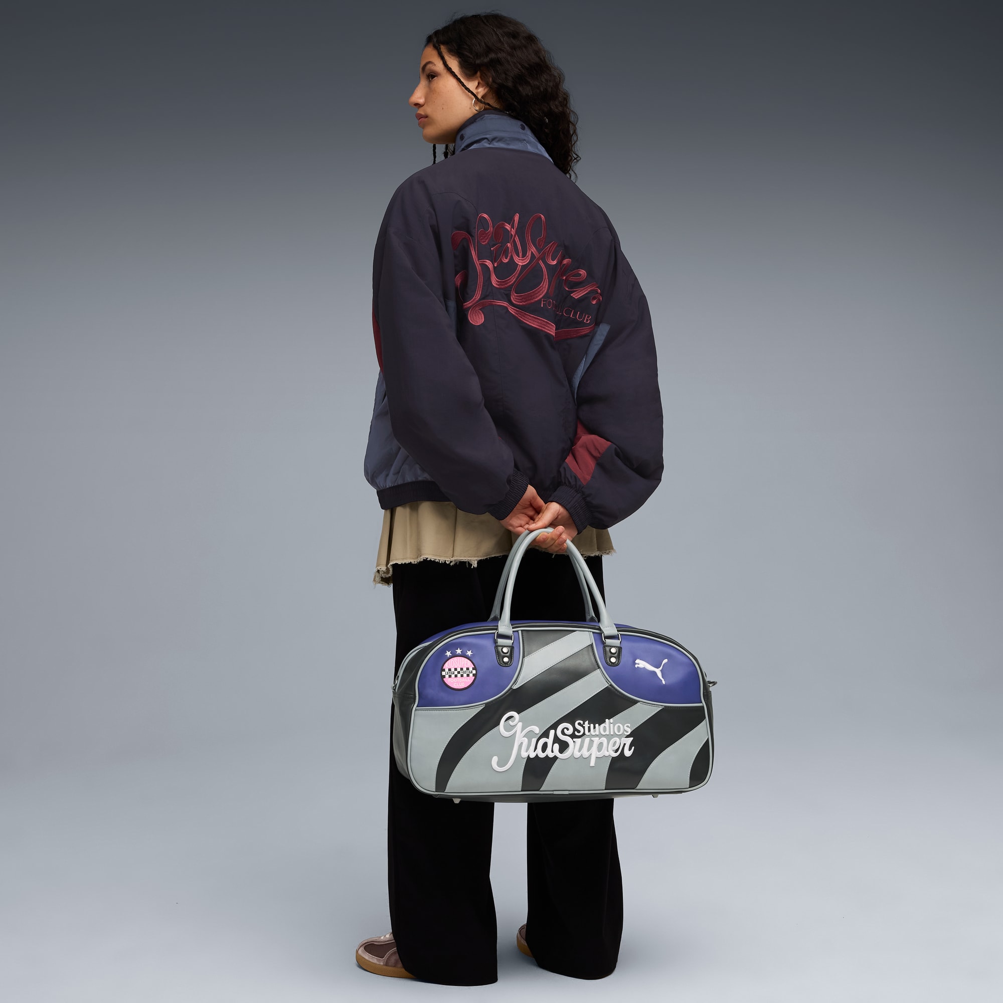 Sur fond gris, un mannequin tient un sac, porte une veste et un pantalon, regardant à gauche.
