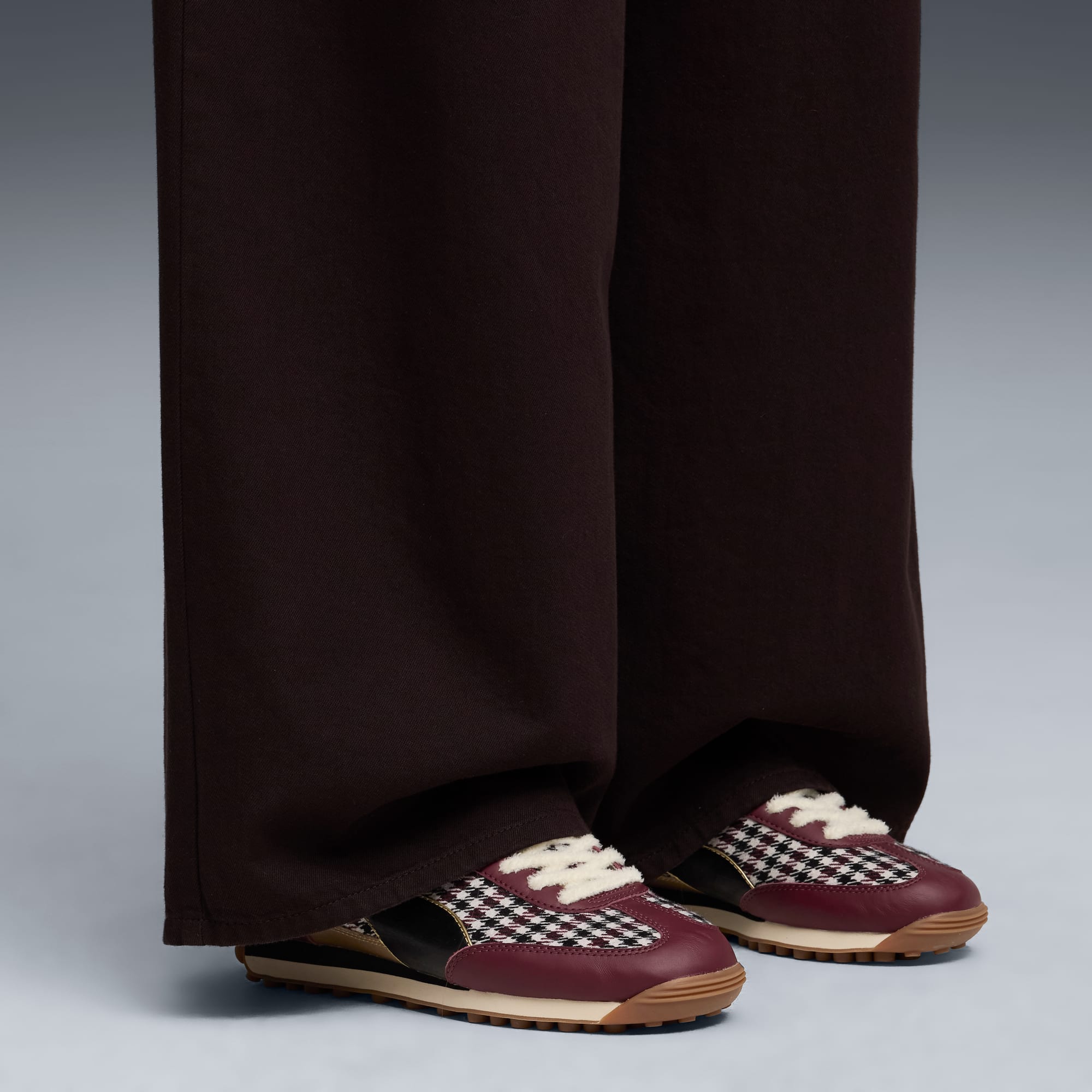 Tegen een grijze achtergrond draagt een model een bruine broek en bordeauxrode sneakers met patroon.