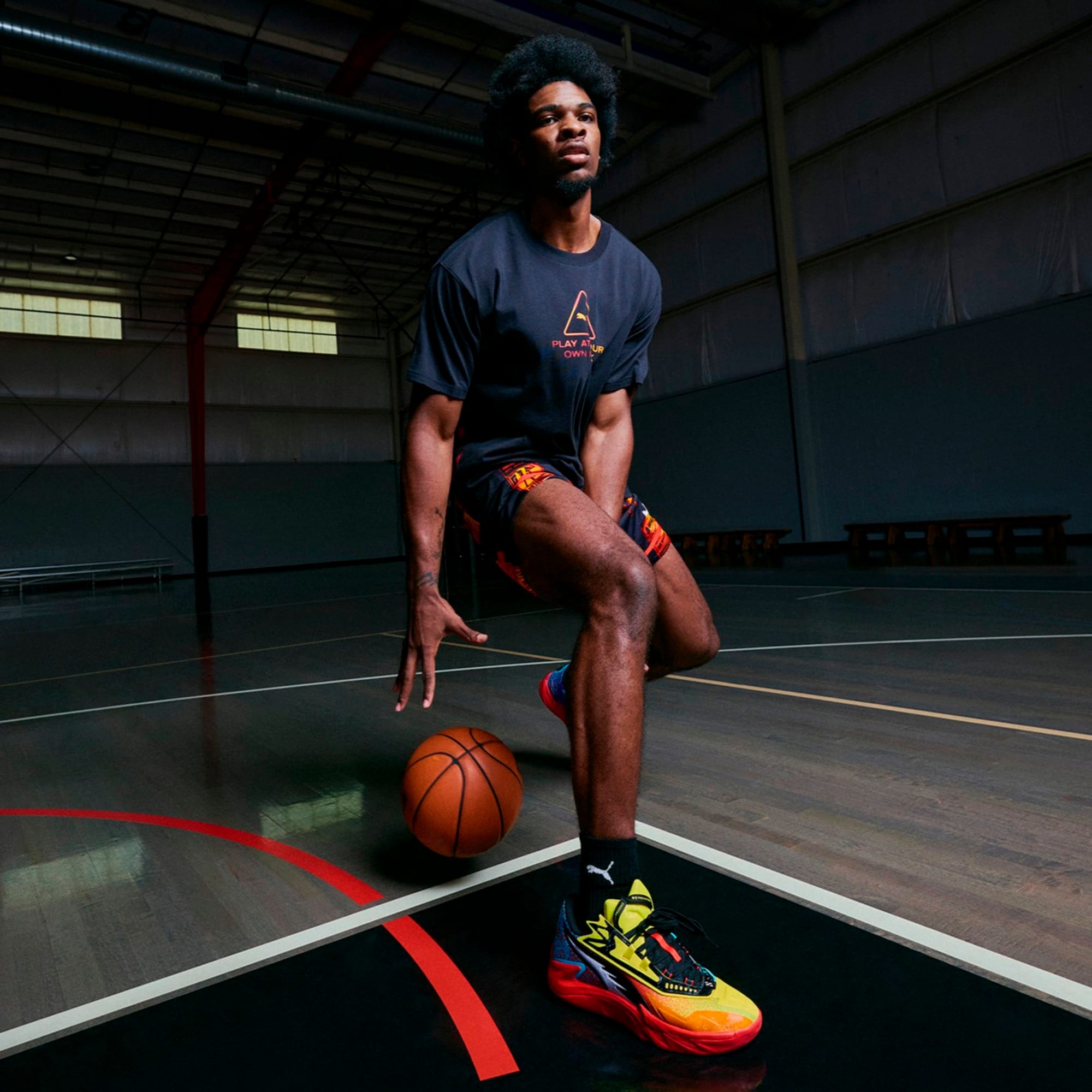 Su un campo, un modello palleggia un pallone, indossando una maglietta scura, pantaloncini fantasia e scarpe colorate.