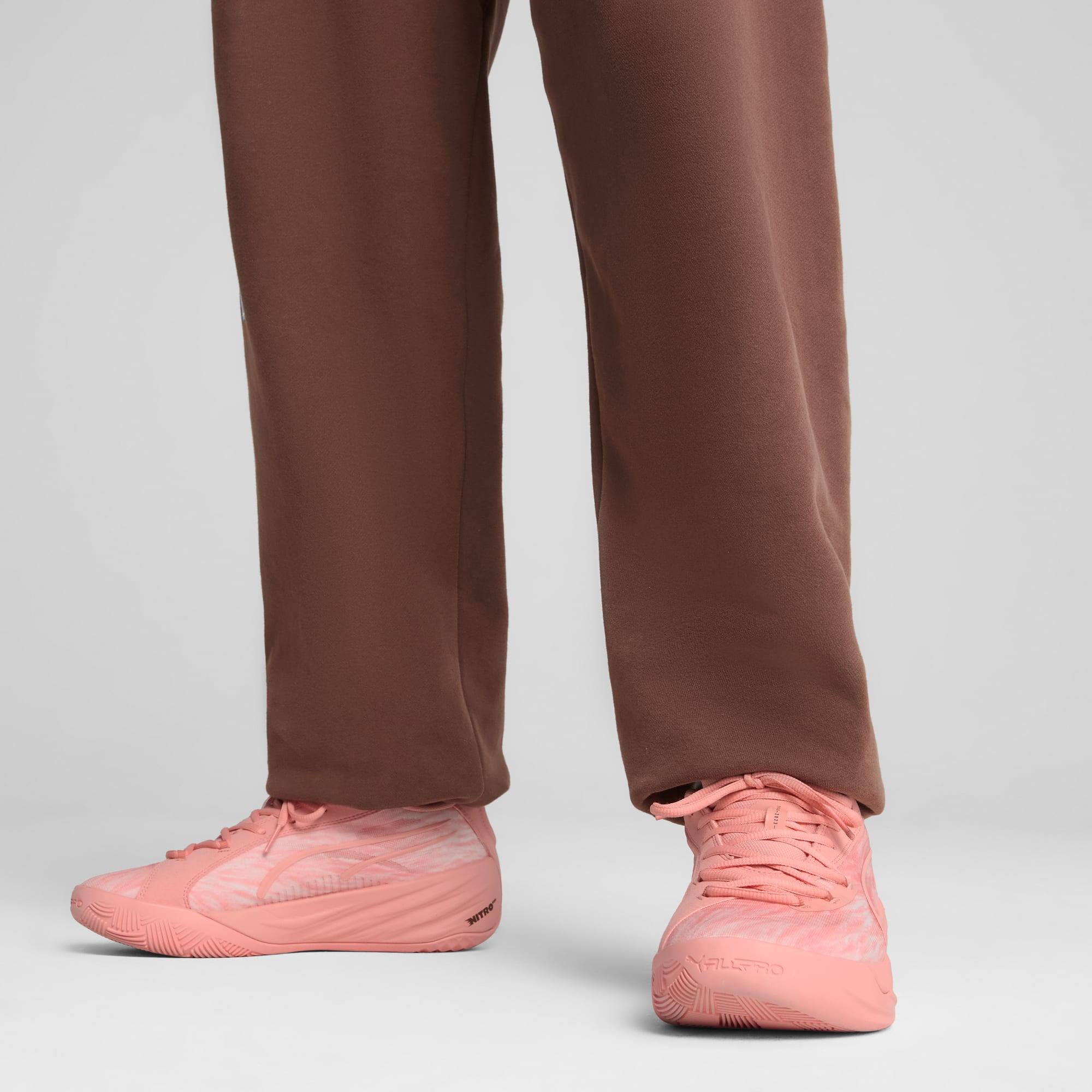 Vor grauem Hintergrund trägt ein Model rosafarbene Turnschuhe und braune Hosen und steht.