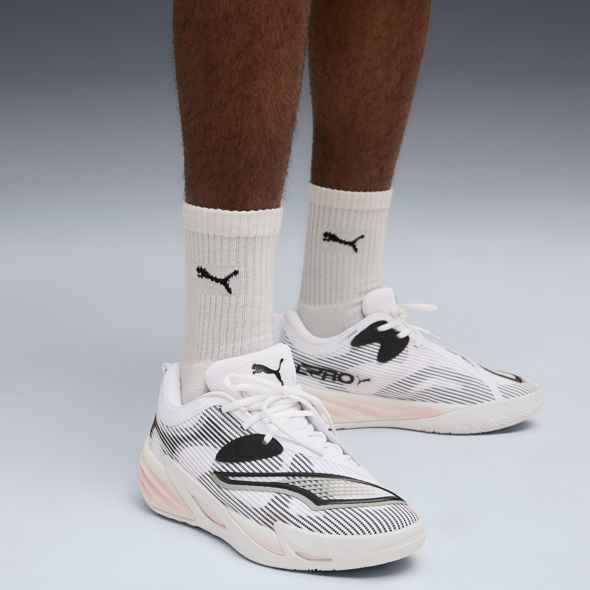 Une personne se tient sur un fond gris, portant des baskets blanches à motifs et des chaussettes à logo.