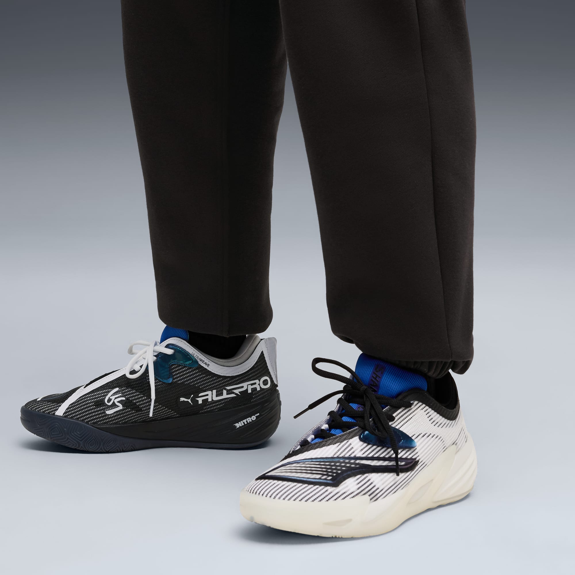 Su uno sfondo grigio, una persona indossa pantaloni da jogging scuri e scarpe da corsa di colori diversi.