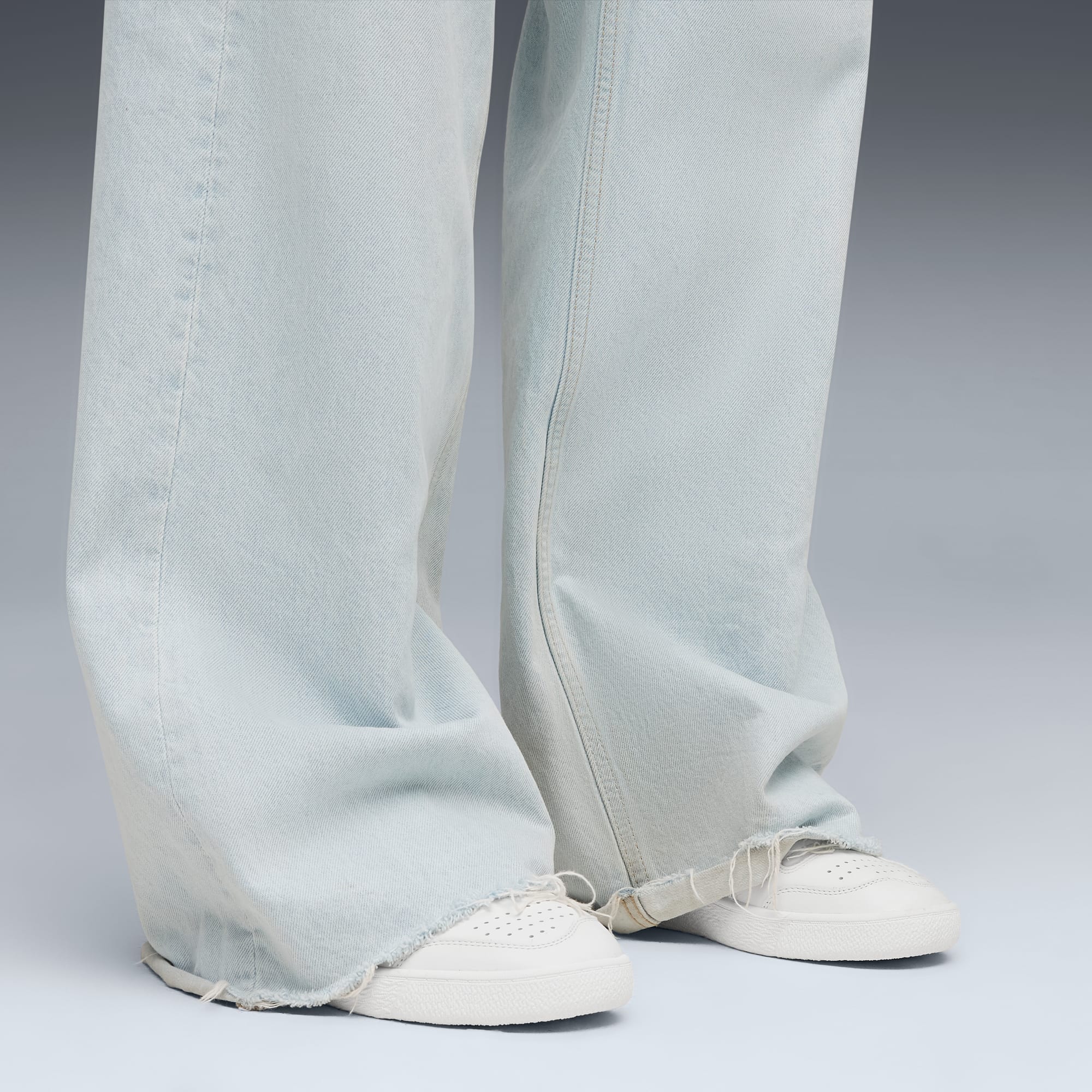 Tegen een grijze achtergrond draagt iemand een lichtblauwe jeans en witte sneakers.