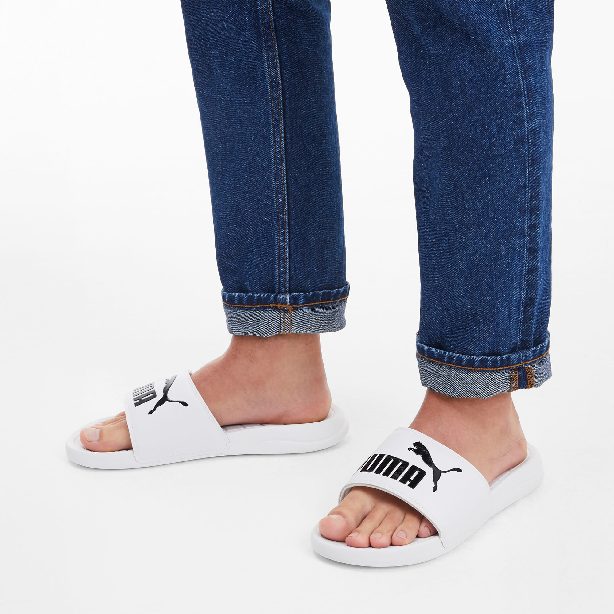 Auf einer weißen Fläche trägt eine Person Jeans und weiße Pantoletten mit Puma-Logo.