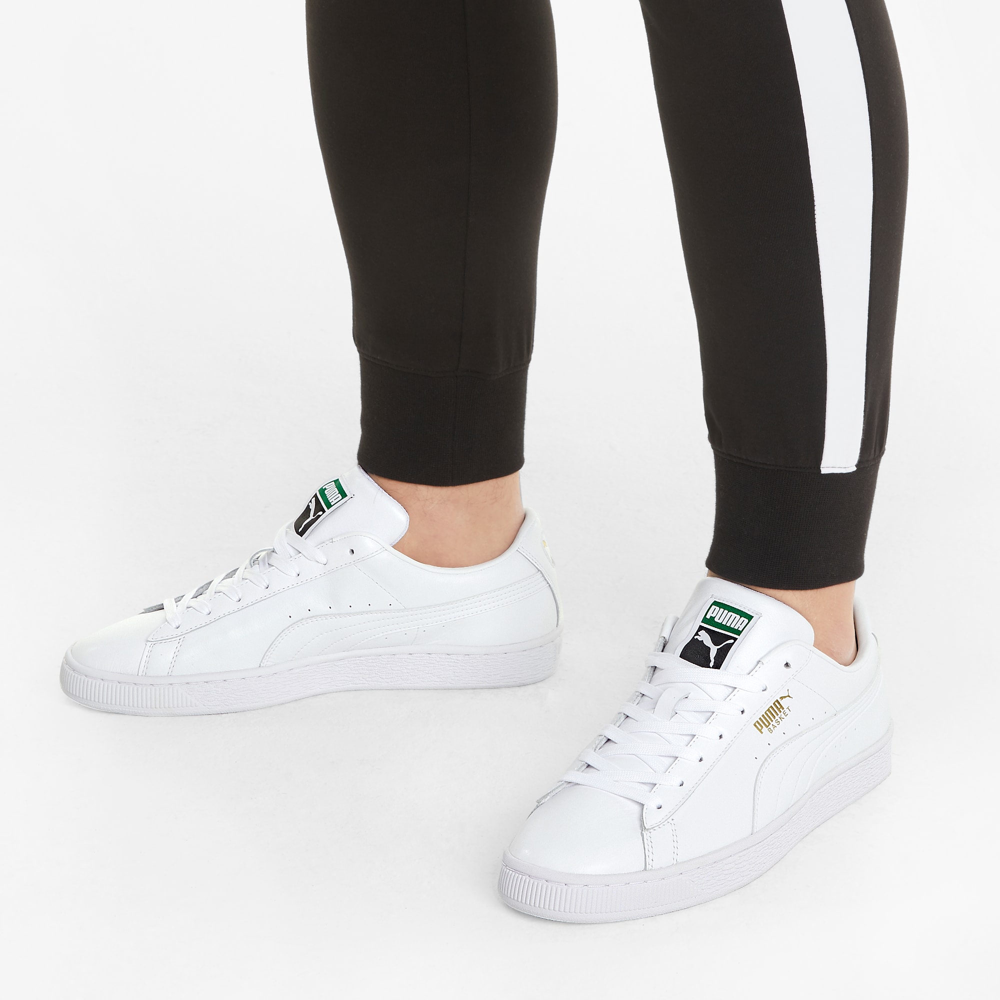 Sobre blanco, una persona usa joggers negros y zapatillas blancas con logo Puma.