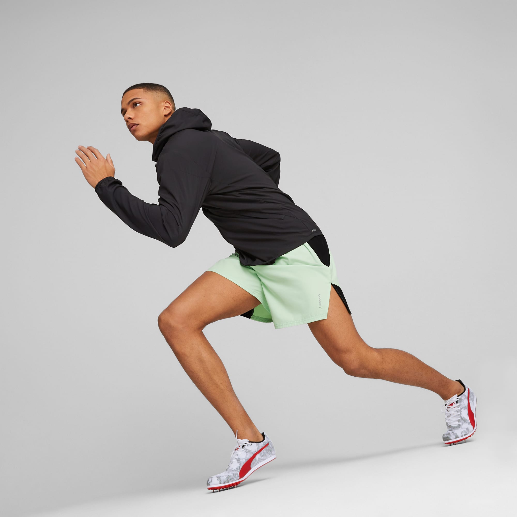 Un modelo en movimiento sobre un fondo gris, con chaqueta negra, pantalón corto verde y zapatillas.