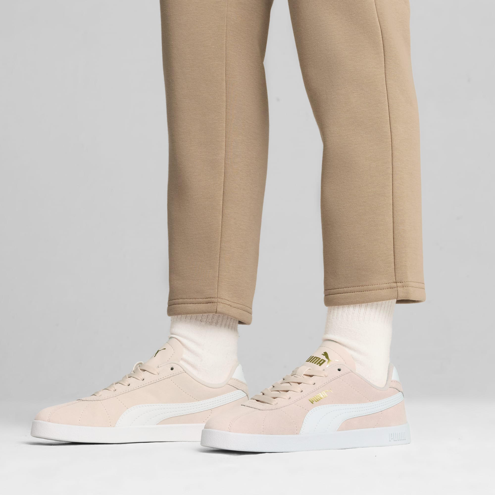 Against a grey backdrop, a model wears beige trousers and light pink trainers.