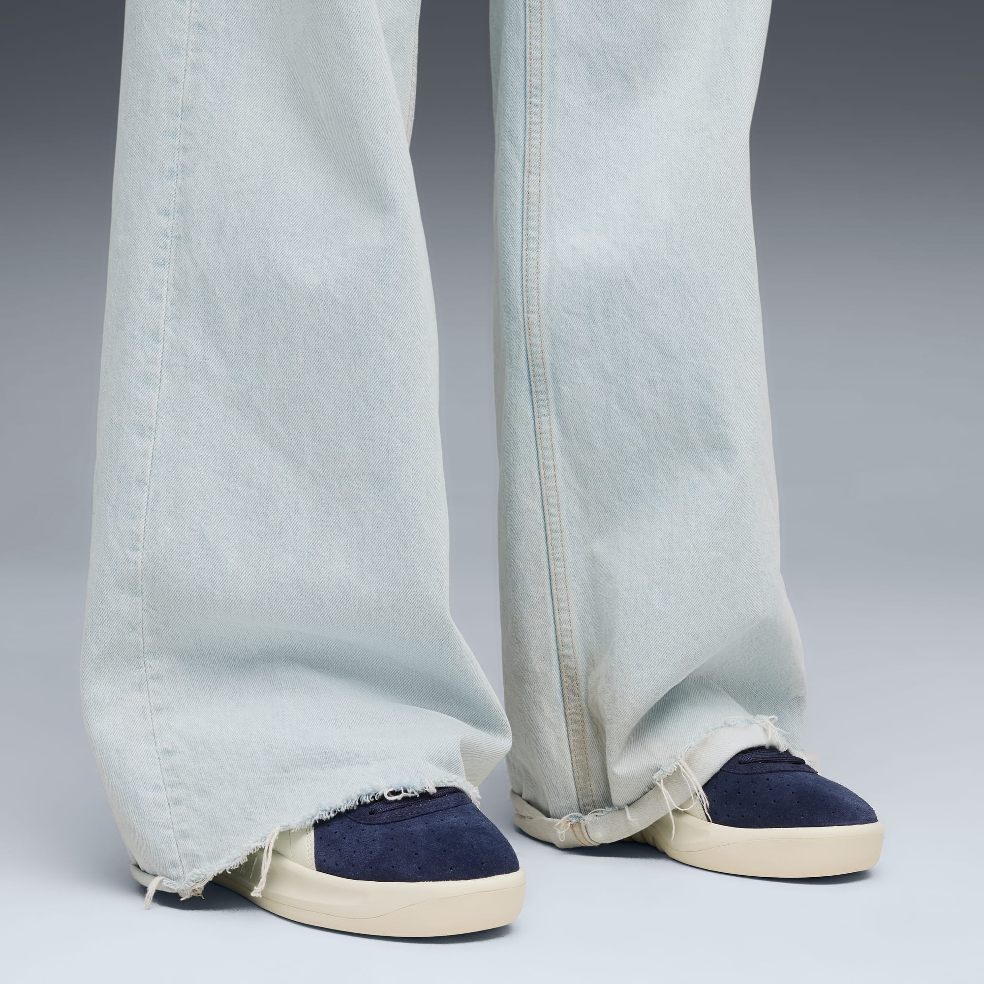 Su sfondo grigio, gambe con jeans larghi chiari e sneakers blu navy e crema.