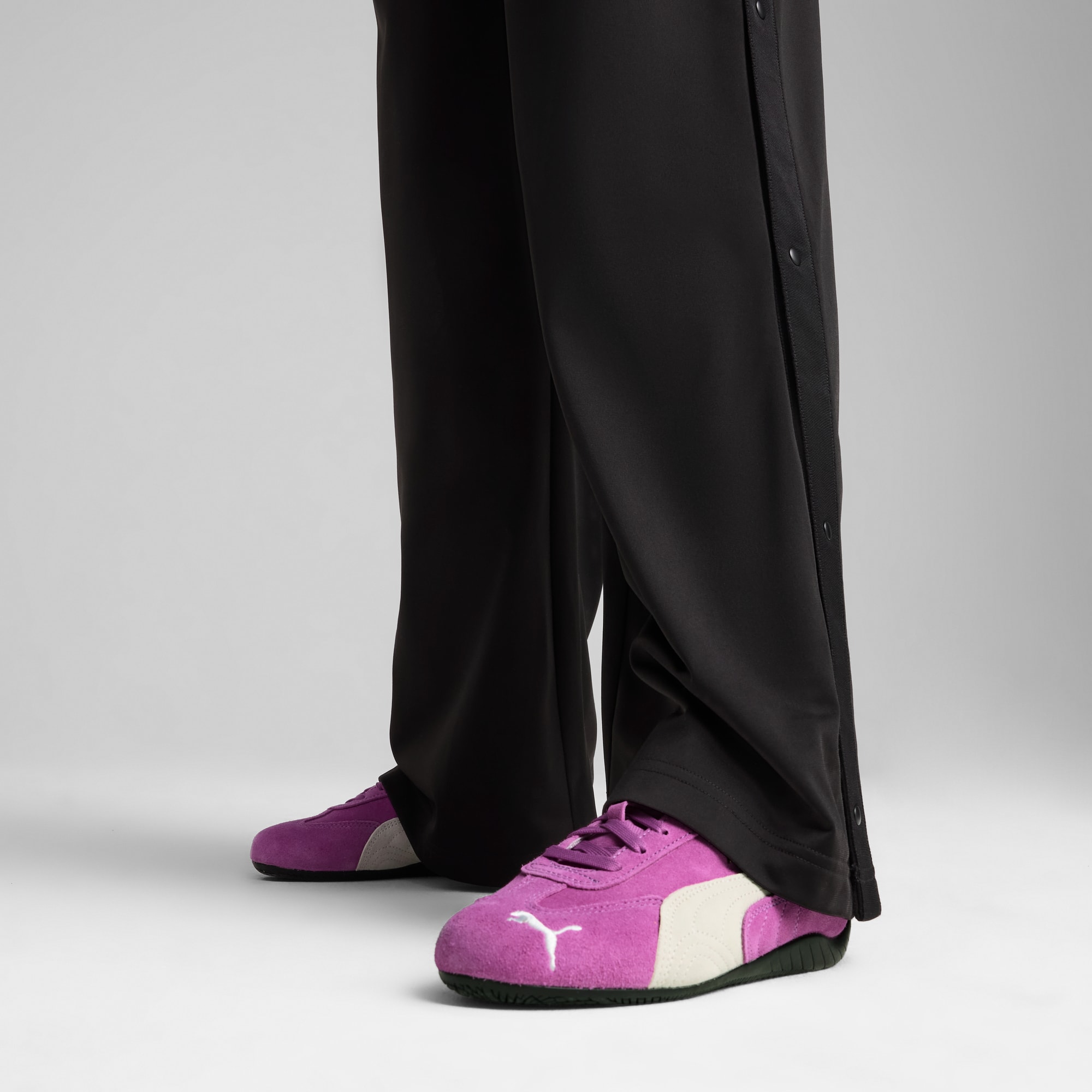 Sur fond gris, un modèle porte un pantalon noir à boutons-pression et des baskets violettes.