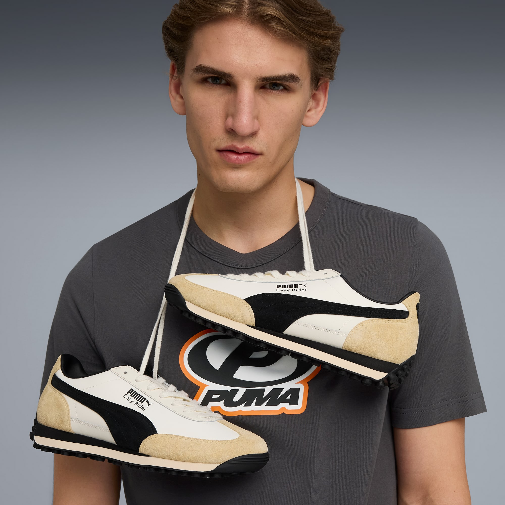 Su sfondo grigio, un modello indossa scarpe da ginnastica Puma al collo, sopra una t-shirt grigia con logo Puma.