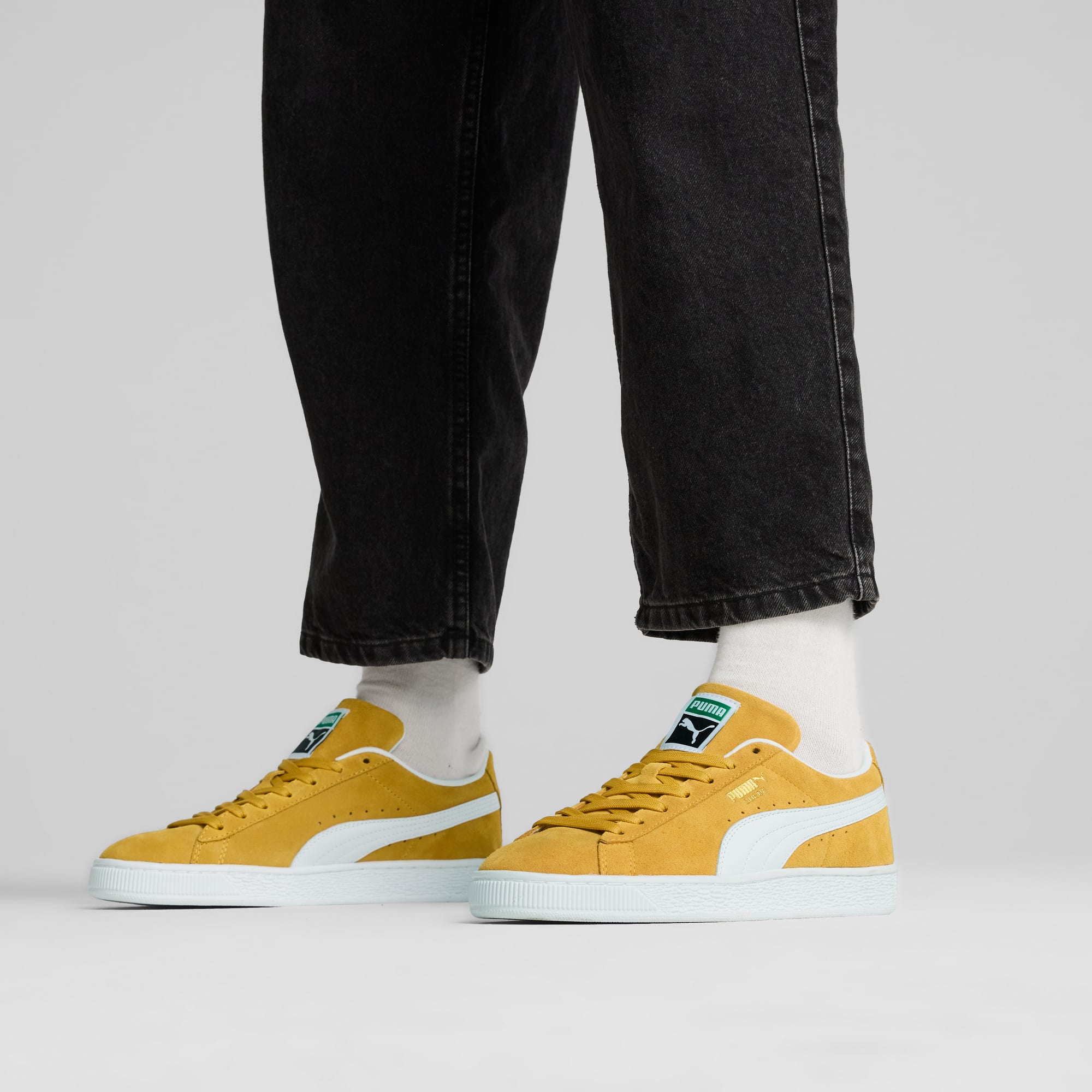 Tegen een grijze achtergrond draagt een model een zwarte jeans met mosterdkleurige sneakers en witte sokken.