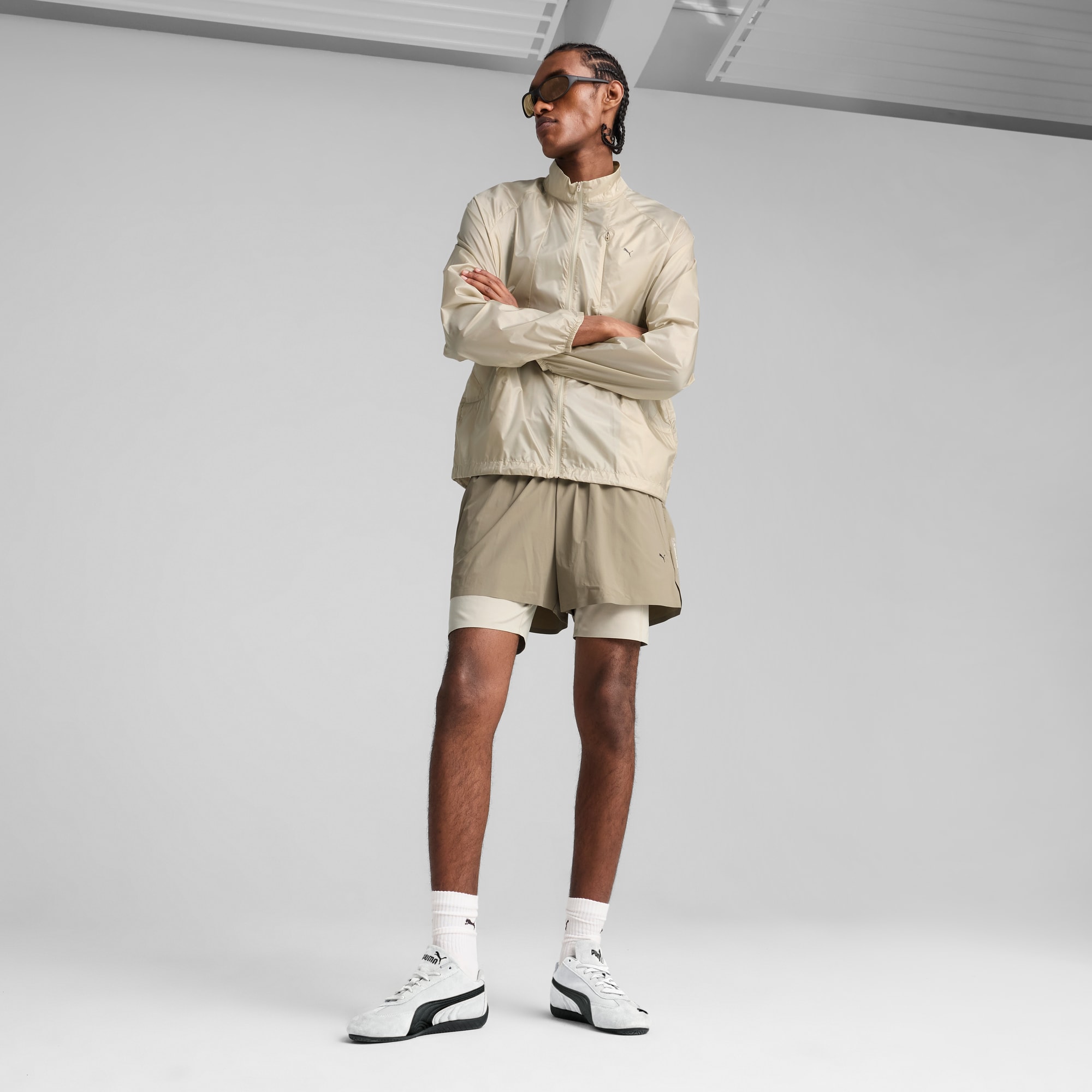 Een model met een beige jas en korte broek staat met gekruiste armen tegen een grijze achtergrond.