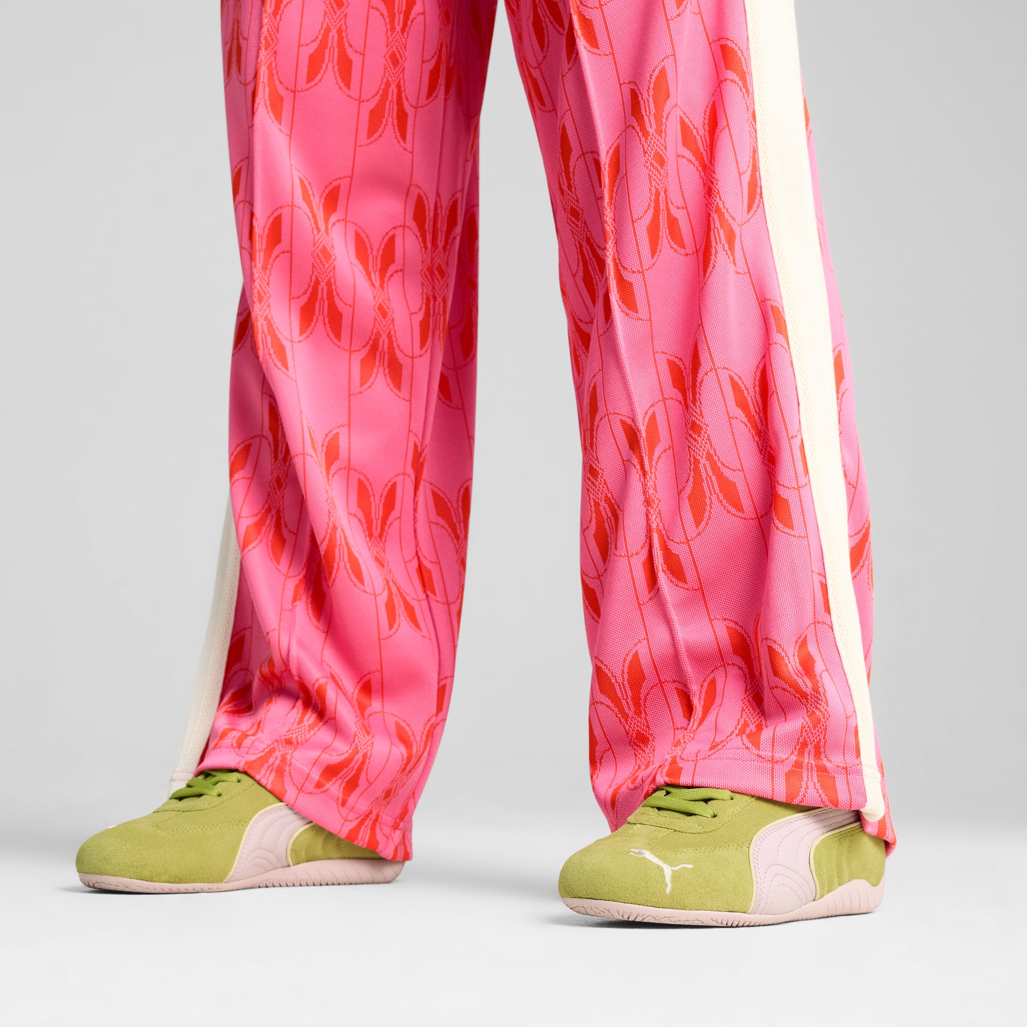 Sur fond gris, une personne porte un pantalon rose à motifs et des baskets vertes.