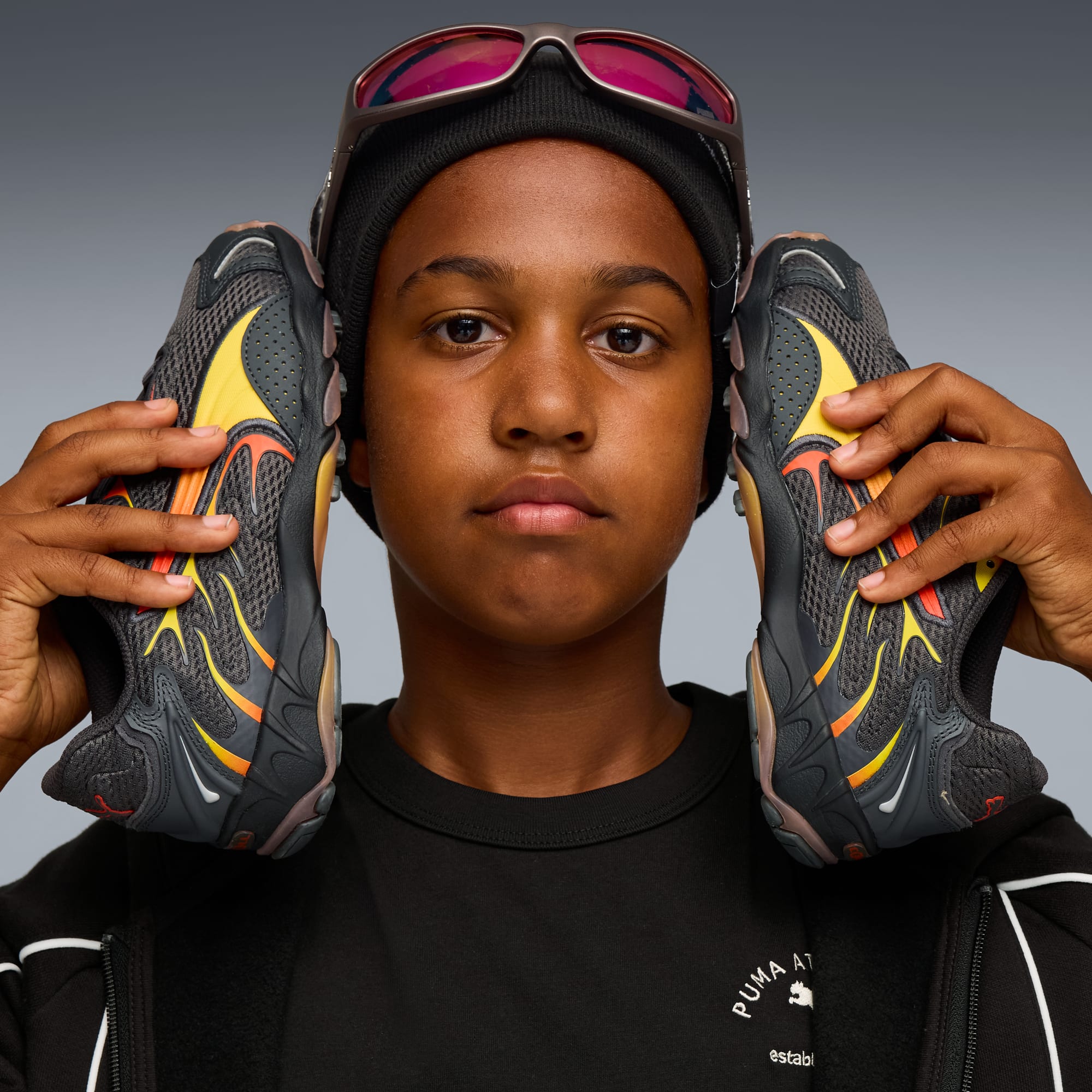 Su sfondo grigio, un modello tiene scarpe da ginnastica grigie, gialle e rosse alle orecchie.