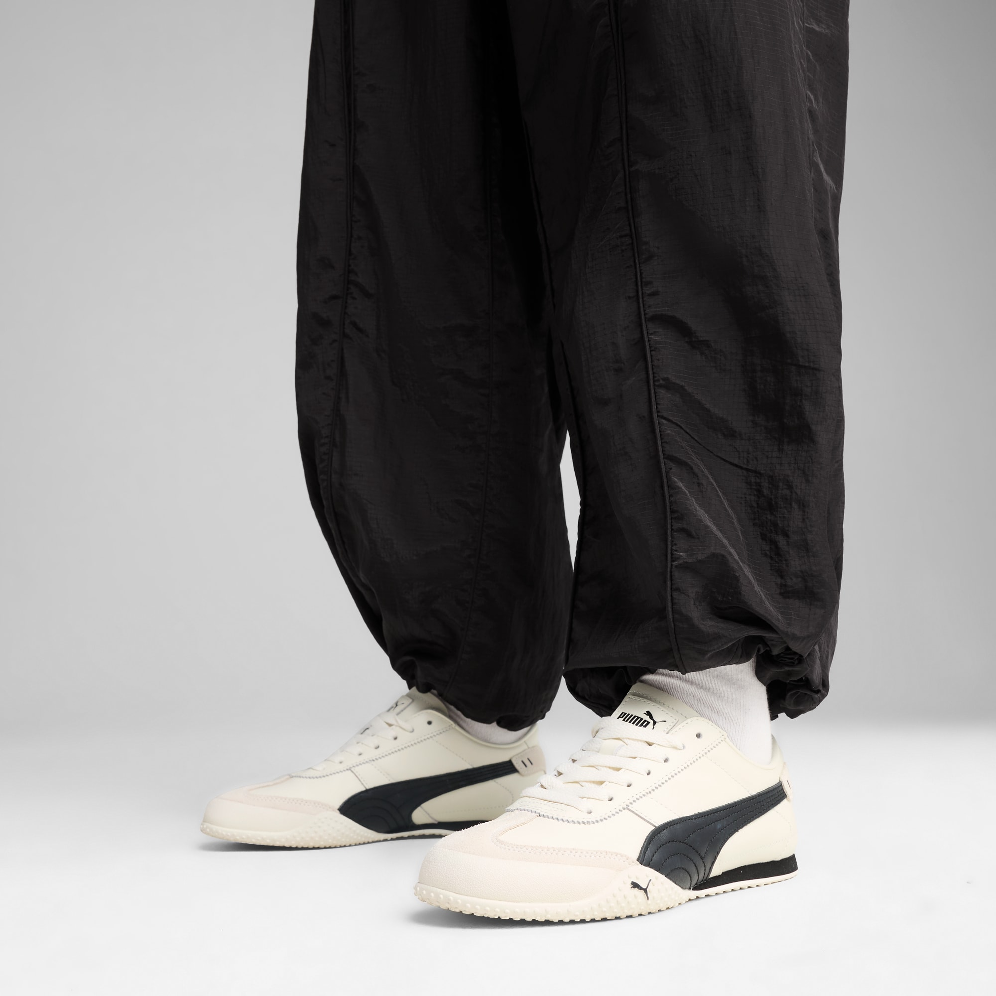 Vor grauem Hintergrund steht ein Model mit schwarzer Hose und cremefarbenen Turnschuhen.