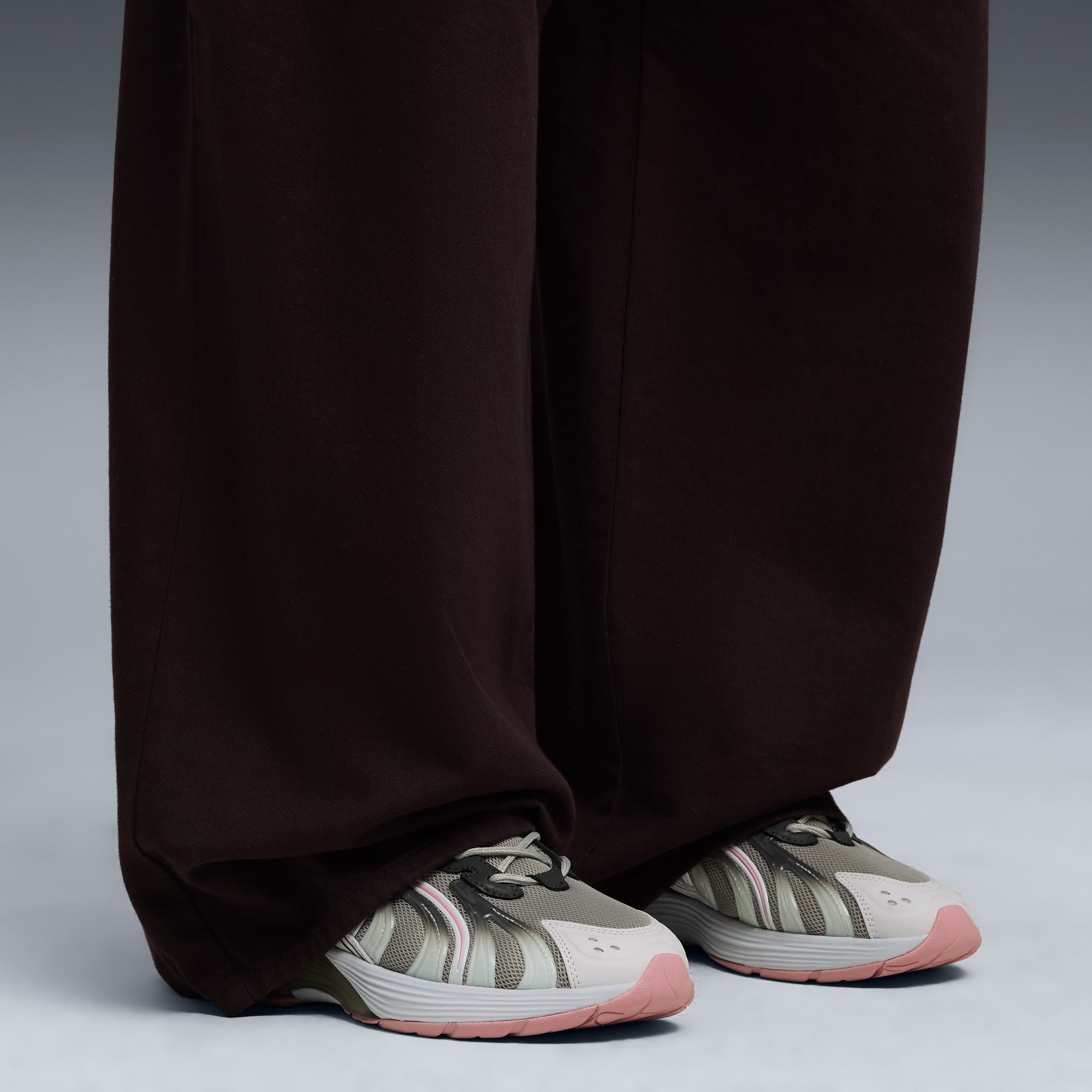 Sur fond gris, un modèle porte un pantalon marron et des baskets grises à semelles roses.