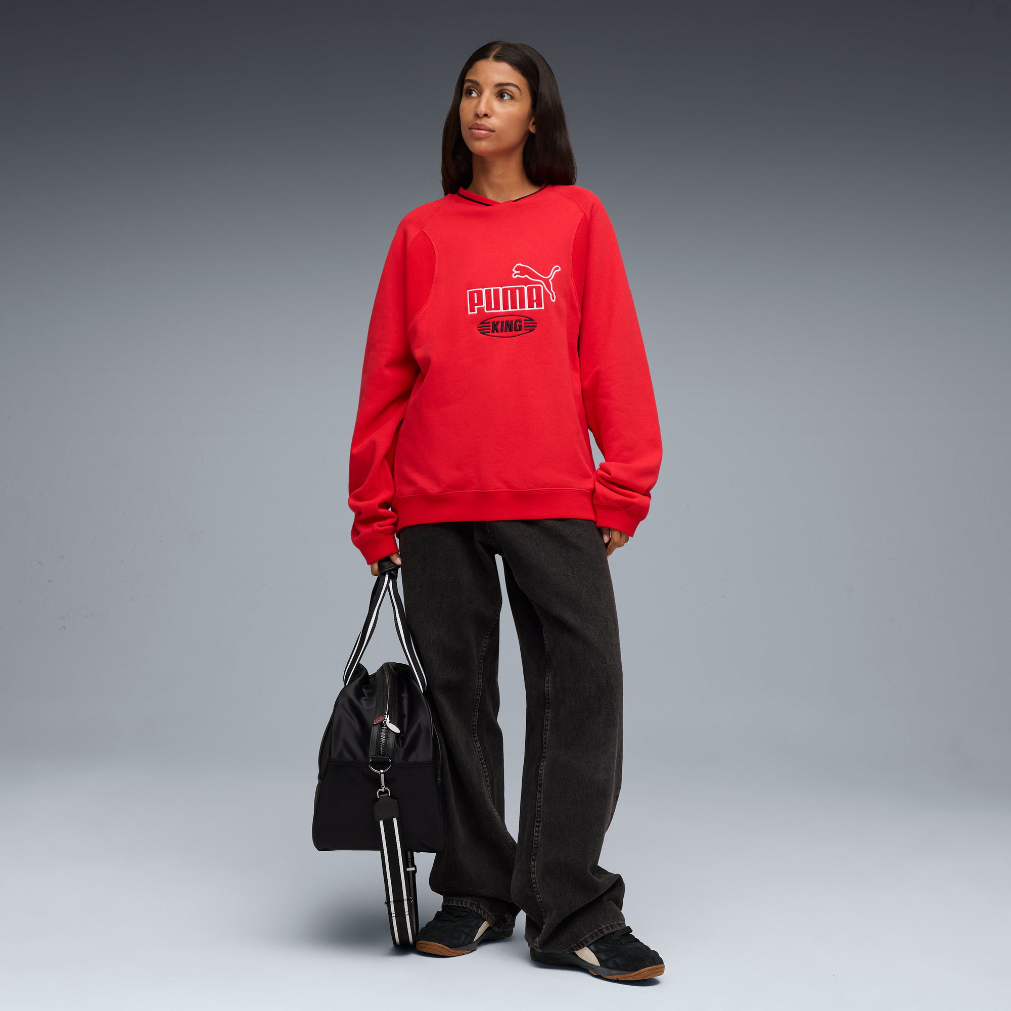 Su sfondo grigio, modella in felpa Puma rossa, jeans scuri e borsa nera.