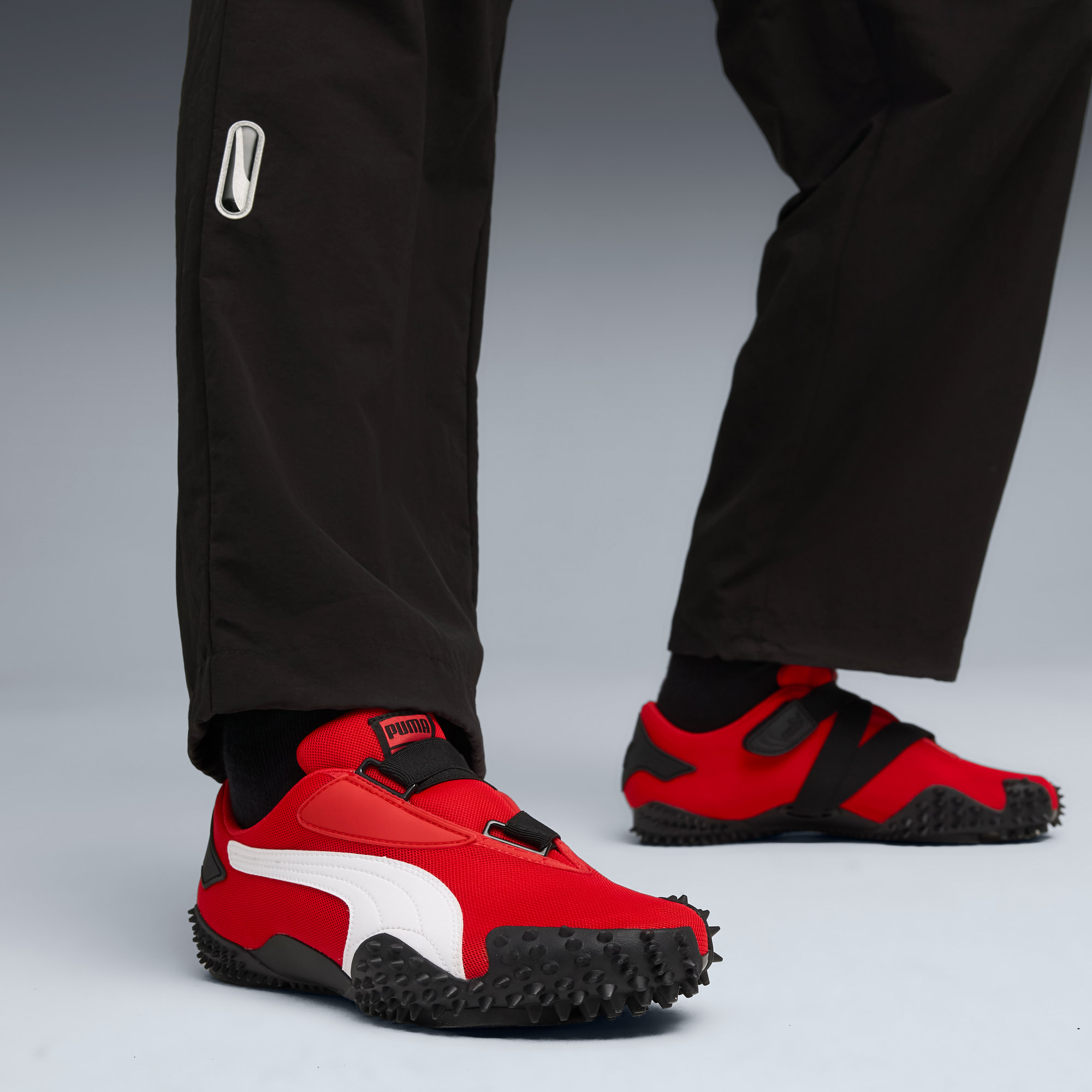 Sur fond gris, un modèle porte un pantalon noir et des baskets rouges à bande blanche.