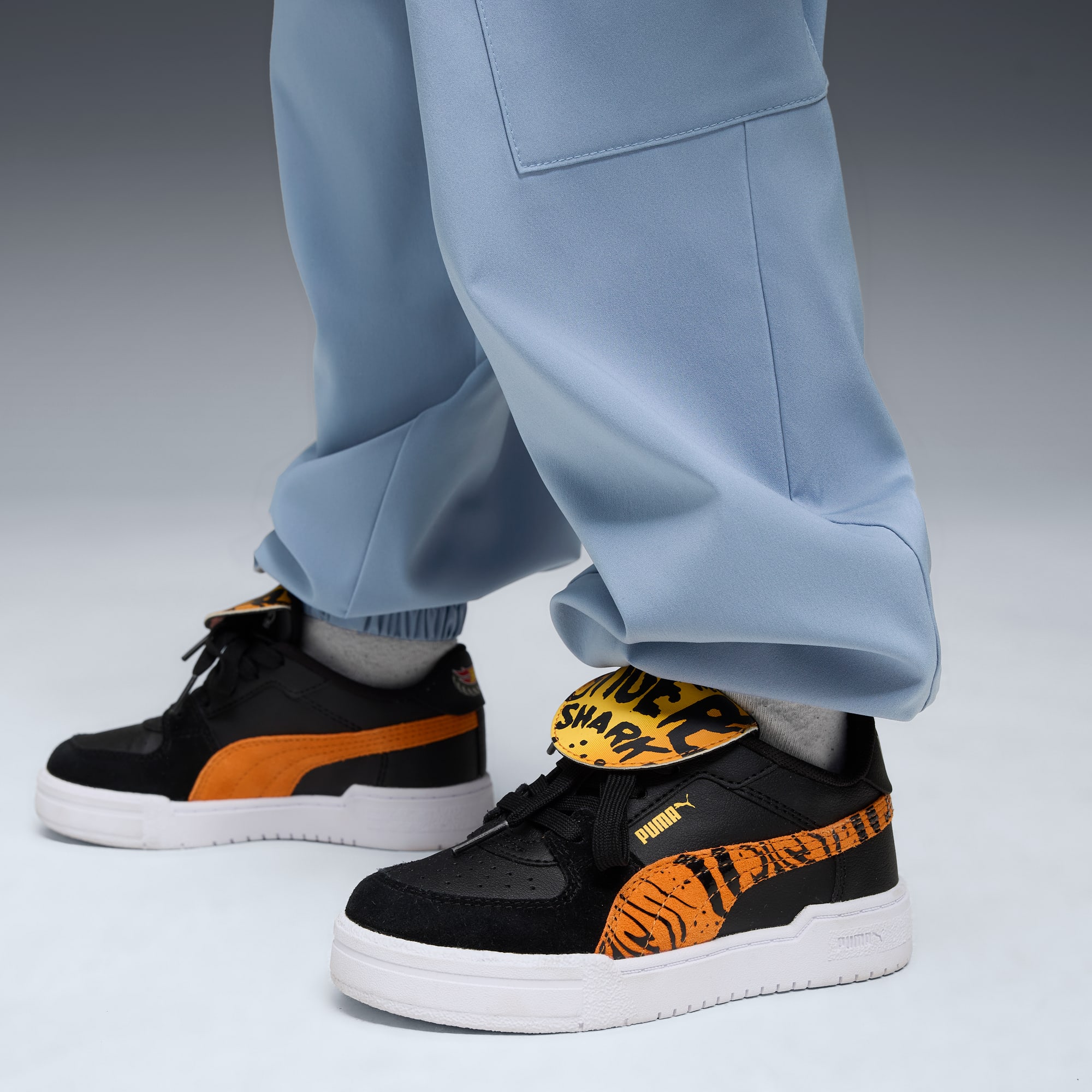 Tegen een grijze achtergrond draagt een model een lichtblauwe broek en zwart-oranje sneakers.