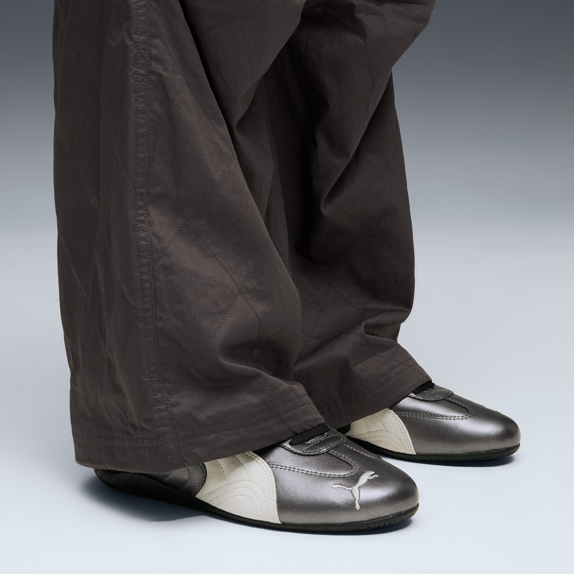 Su sfondo grigio, una persona indossa pantaloni marroni e scarpe da ginnastica Puma metallizzate con strisce bianche.