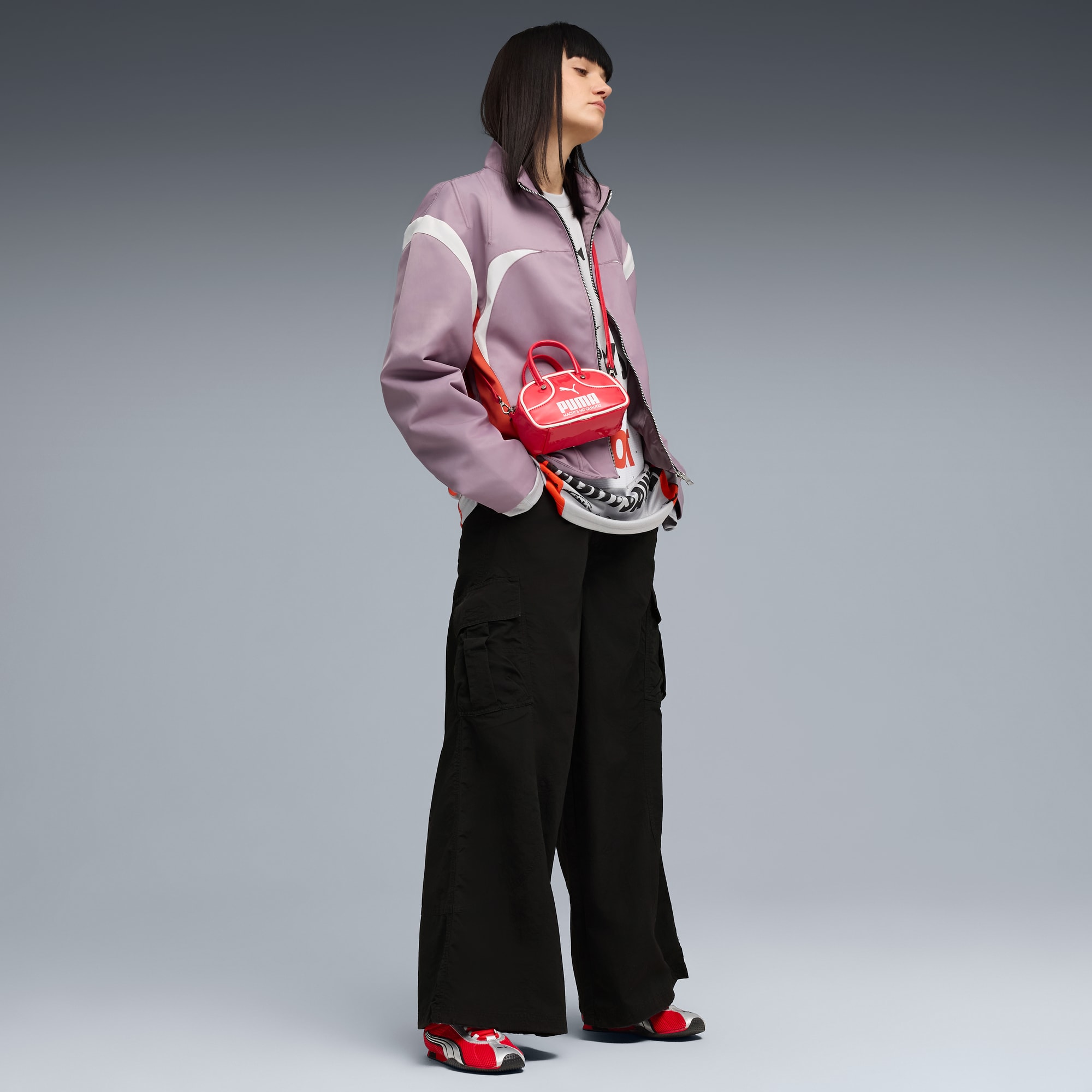 Un modello con giacca lilla, pantaloni neri e borsa rossa posa su sfondo grigio.