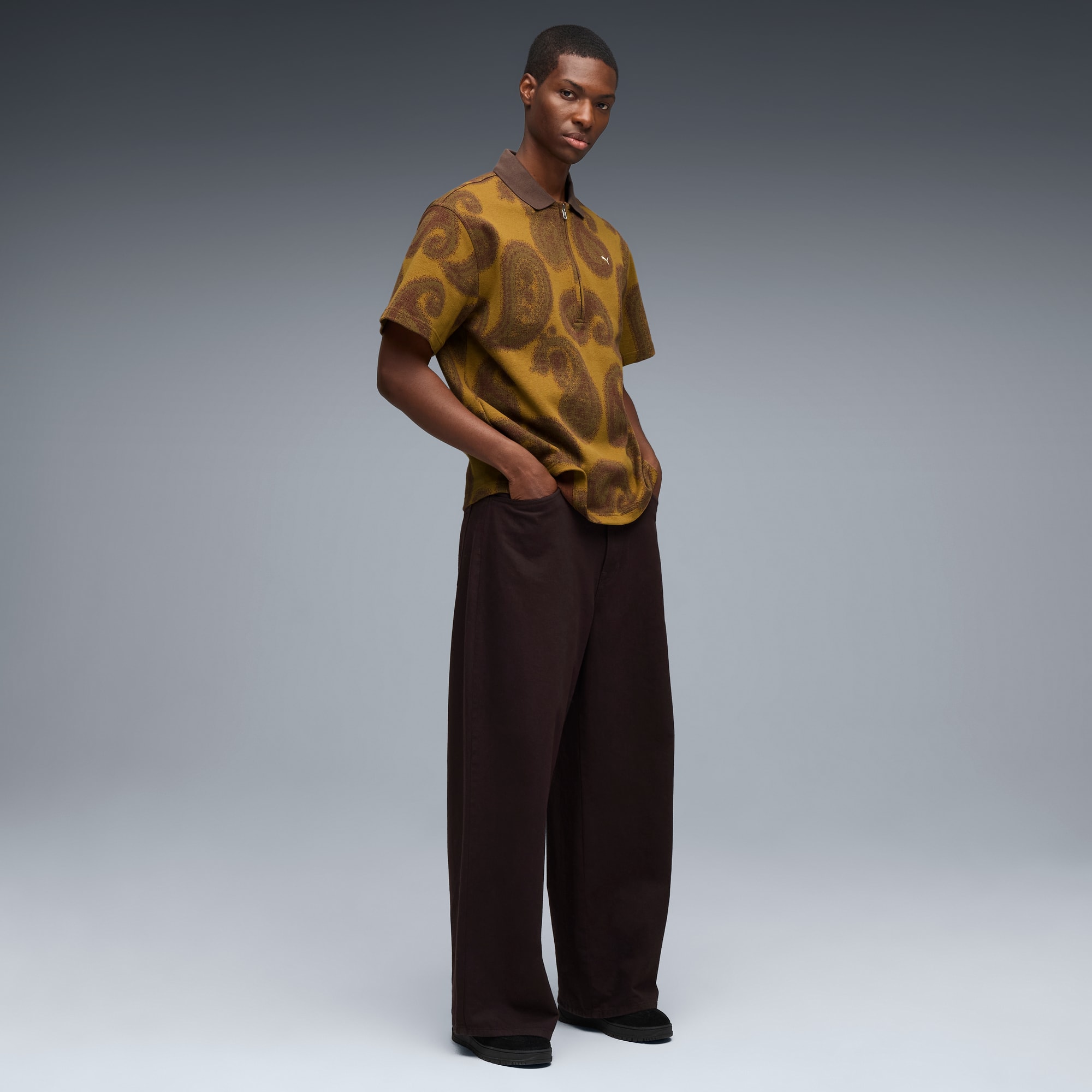 Un modello posa su sfondo grigio con camicia fantasia e pantaloni ampi scuri.