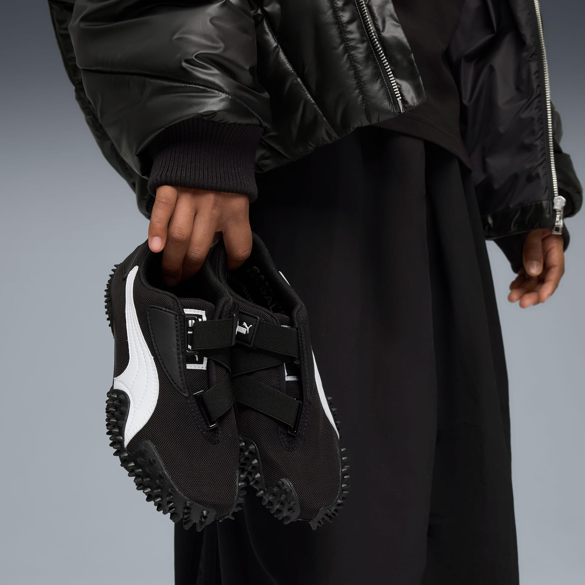 Sur fond gris, une personne tient des chaussures Puma noires à rayures blanches et fermetures.