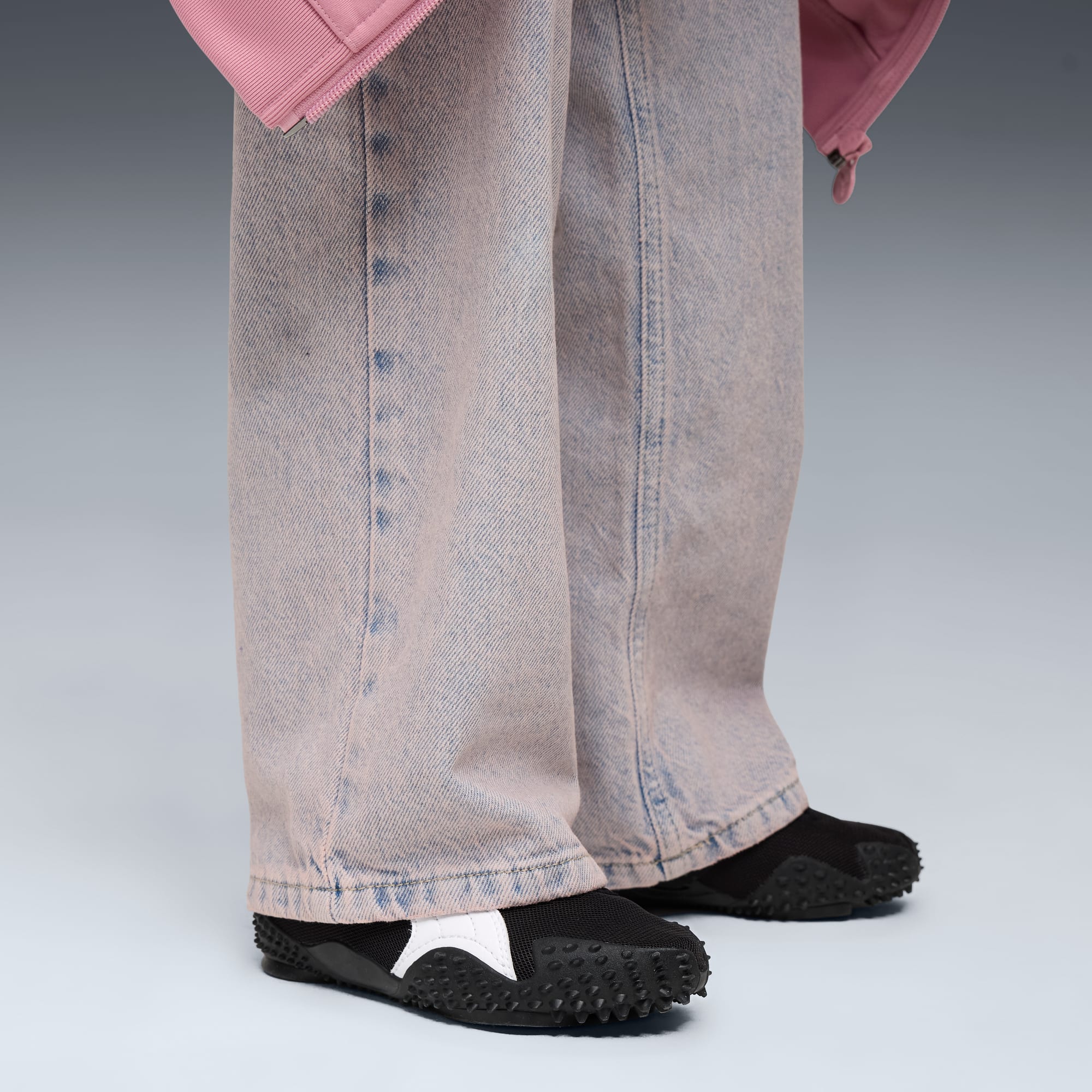 Vor grauem Hintergrund trägt ein Model schwarze Schuhe, helle Jeans und eine rosa Jacke.